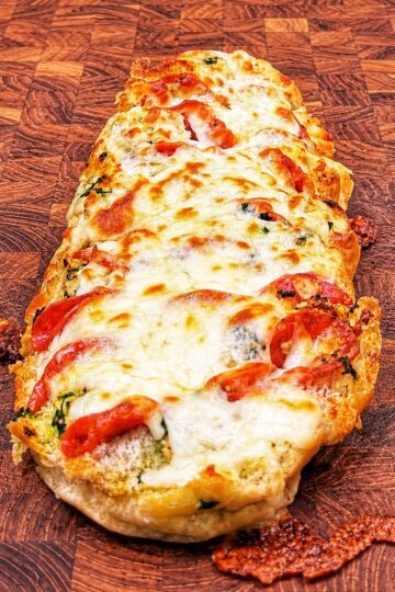 A cheesy baked bread loaf topped with melted mozzarella, tomato slices, and herbs, displayed on a checkered wooden cutting board.