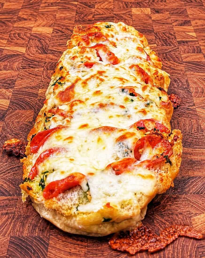 A cheesy baked bread loaf topped with melted mozzarella, tomato slices, and herbs, displayed on a checkered wooden cutting board.