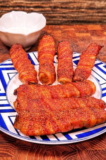 A plate with five bacon-wrapped sausages arranged like fingers and a slab of meat as a palm, resembling a hand, sits on a blue-and-white patterned plate with a white bowl in the background.