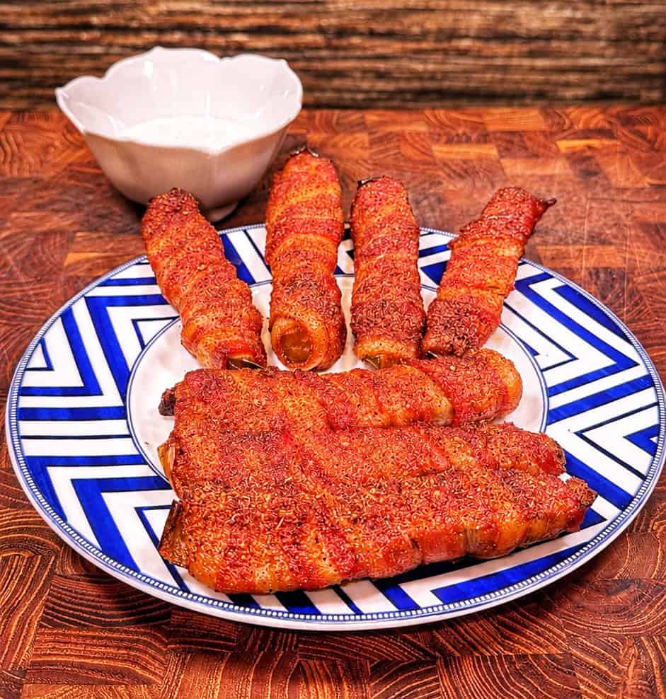 A plate with five bacon-wrapped sausages arranged like fingers and a slab of meat as a palm, resembling a hand, sits on a blue-and-white patterned plate with a white bowl in the background.
