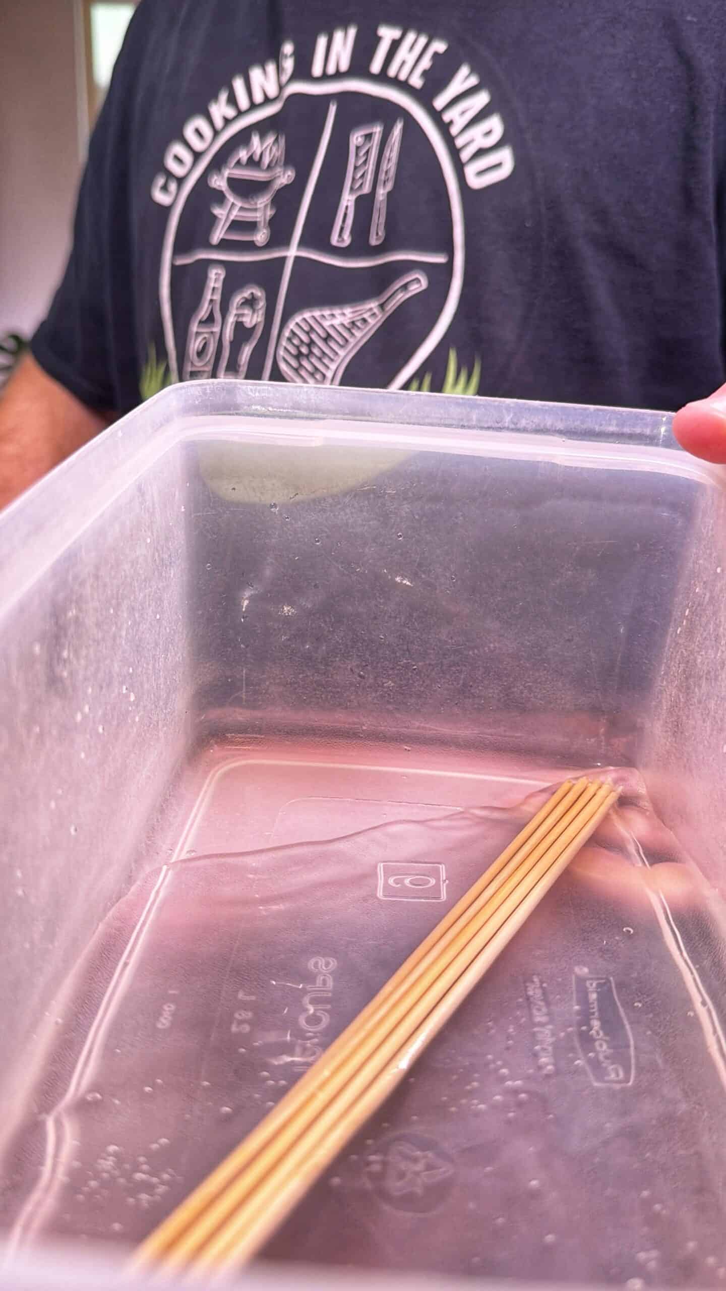 A person wearing a Cooking in the Yard shirt holds a clear plastic container partially filled with water and a few uncooked spaghetti strands inside.