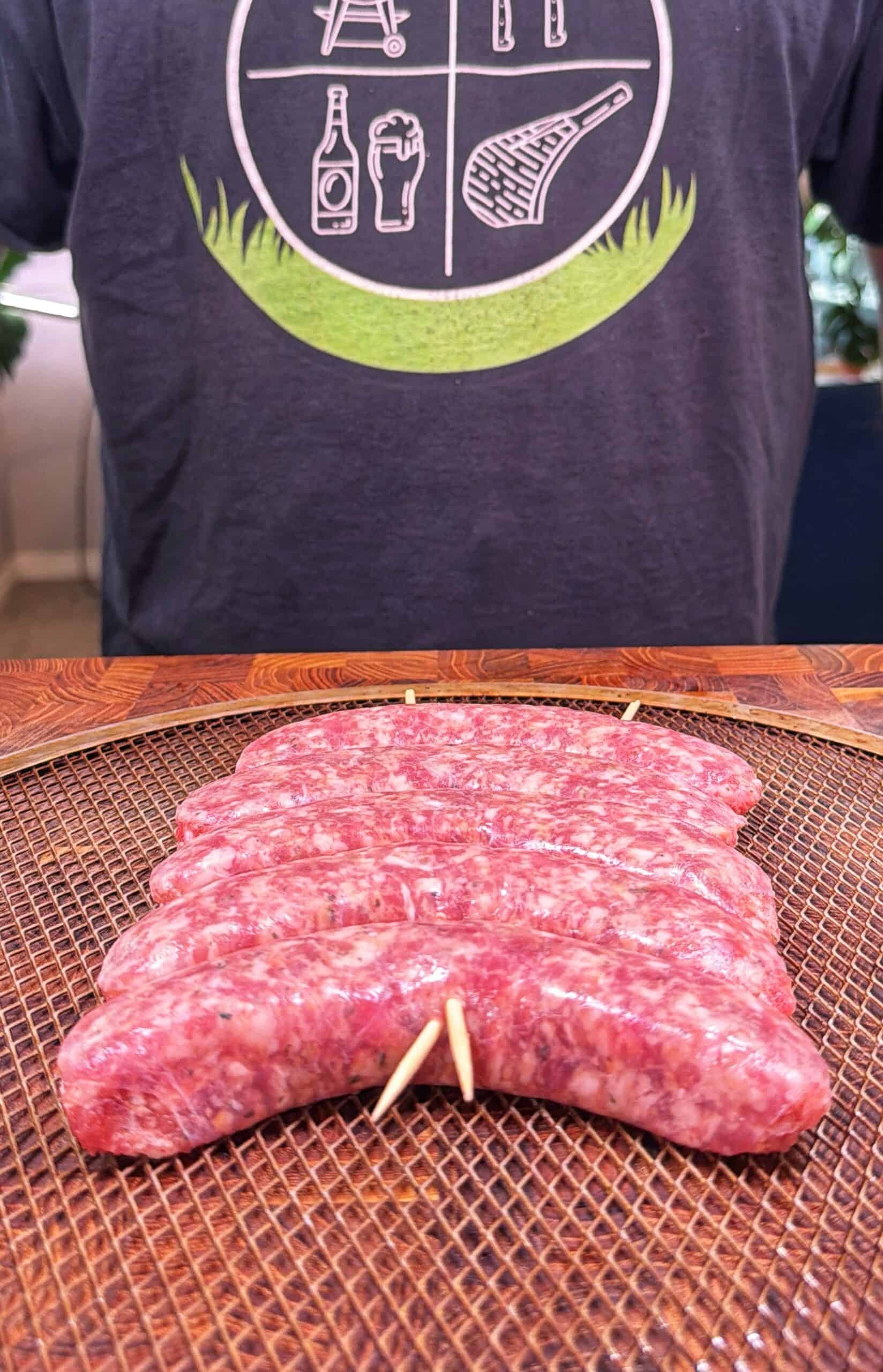 Raw sausage links are arranged side by side with toothpicks holding them together on a wire rack. A person wearing a T-shirt with grill and beer graphics stands behind the sausages.