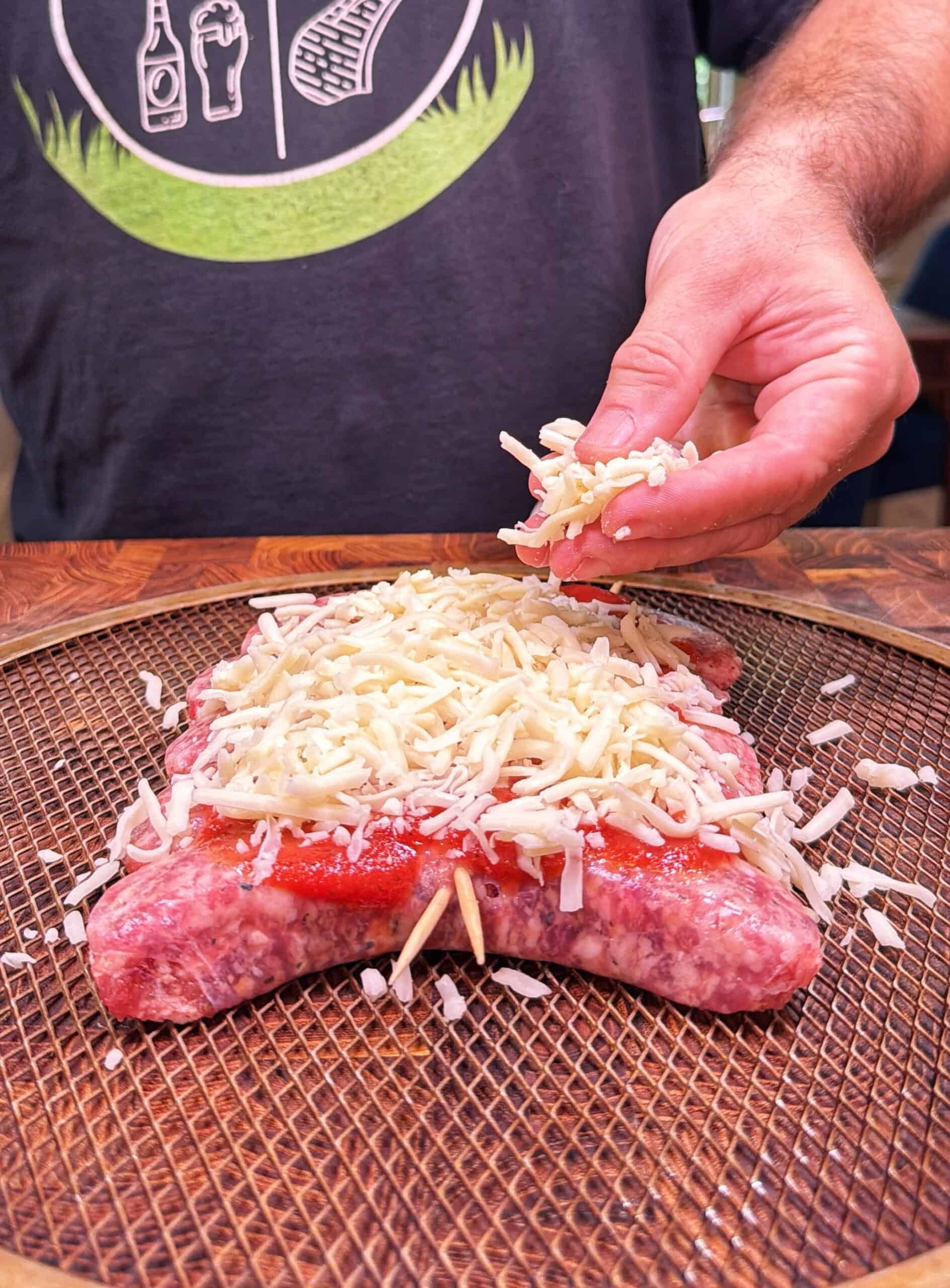 A person sprinkles shredded cheese over raw ground meat shaped like a turtle, topped with sauce, on a wire rack. The person is wearing a dark t-shirt with white designs.
