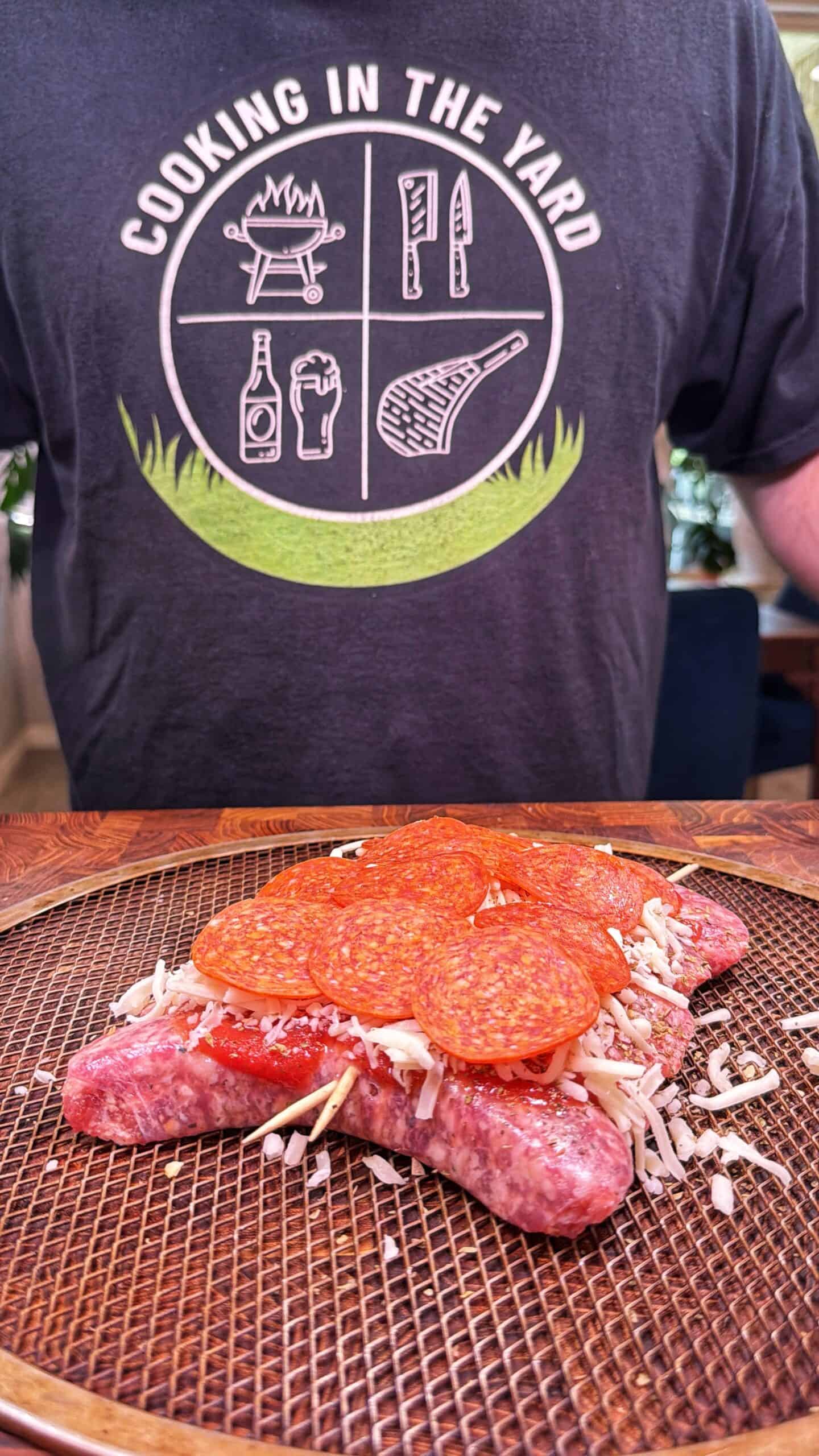 A person wearing a Cooking in the Yard t-shirt stands behind a sausage split open and topped with cheese and pepperoni slices, prepared on a wire grill rack.