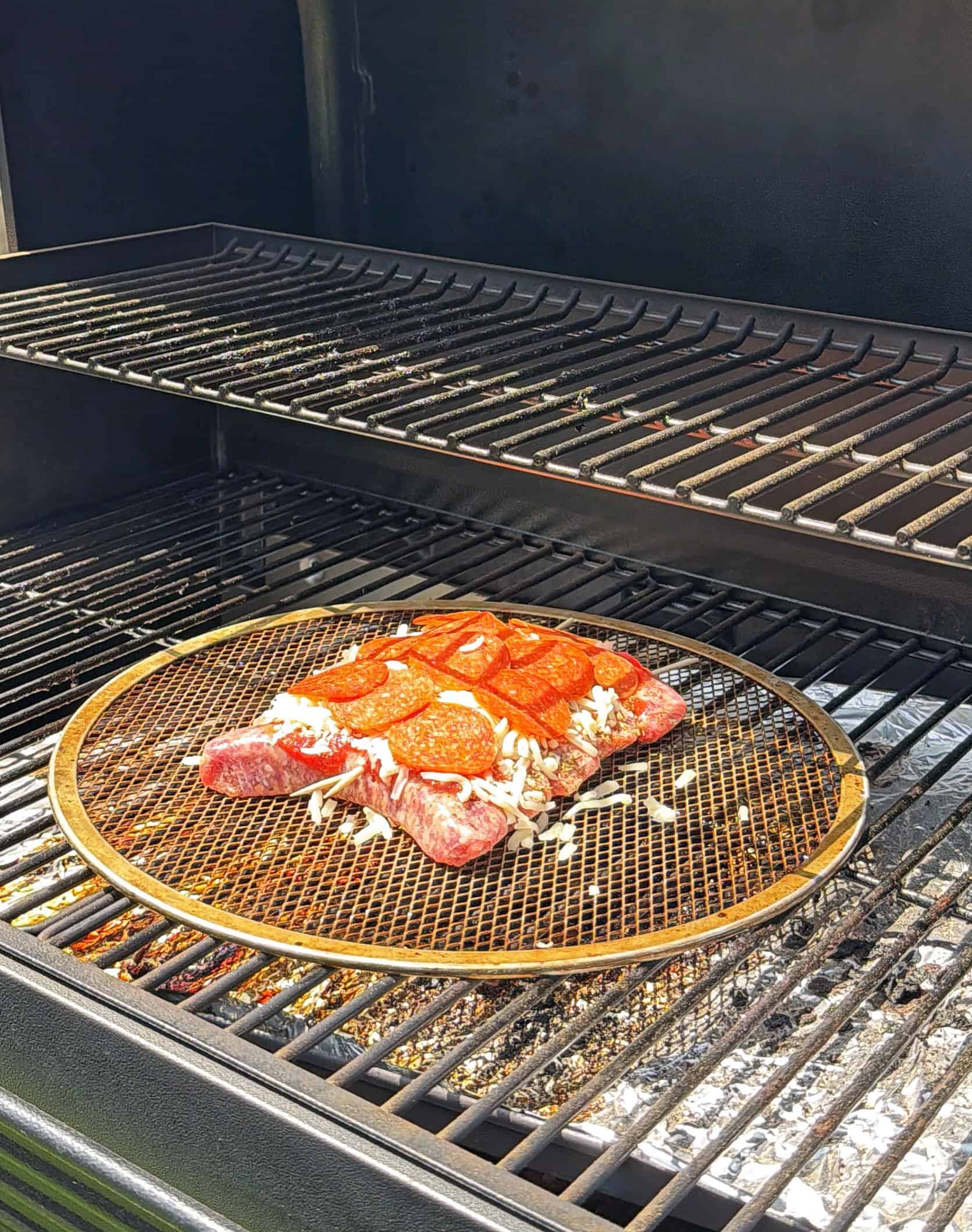 A piece of raw steak topped with shredded cheese and pepperoni slices is placed on a mesh tray inside a barbecue grill, with the grill lid open and sunlight shining on the food.