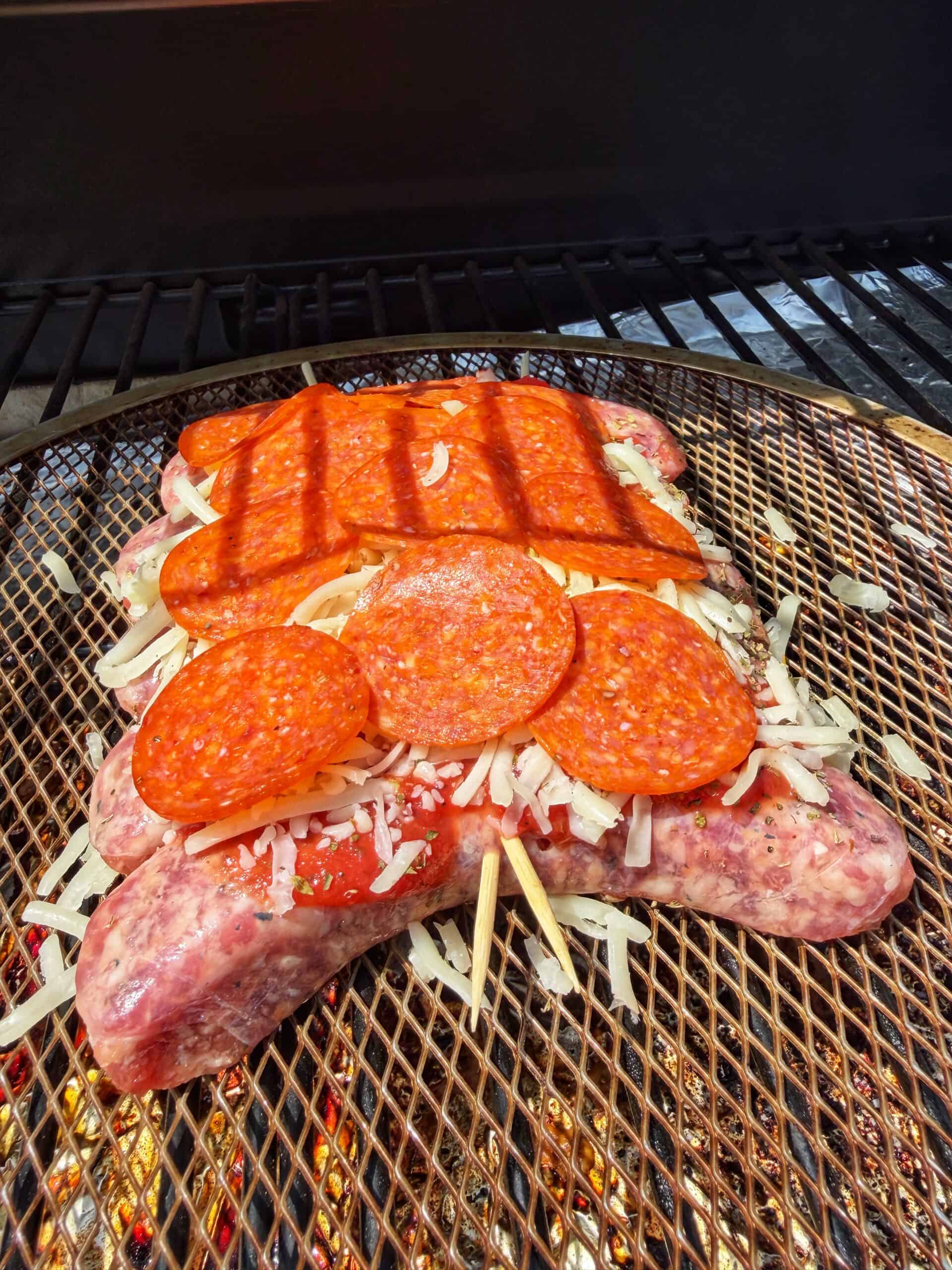 Raw sausages topped with shredded cheese and pepperoni slices are being grilled on a metal mesh rack over open flames. The sausages and toppings are starting to cook, with grill marks visible.