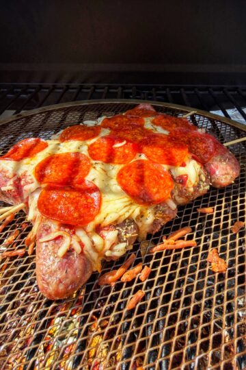 A grilled dish shaped like a pizza slice, made of sausages as the crust, topped with melted cheese and pepperoni slices, placed on a wire rack over a grill.