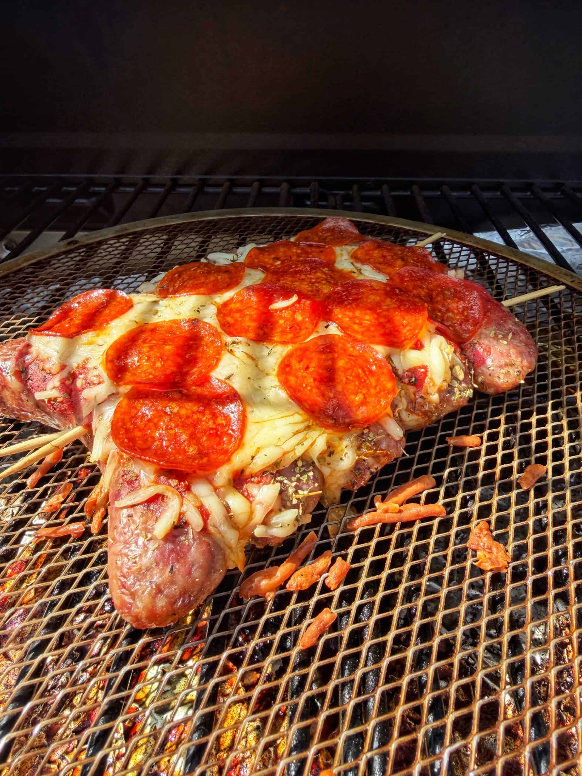 A grilled dish shaped like a pizza slice, made of sausages as the crust, topped with melted cheese and pepperoni slices, placed on a wire rack over a grill.