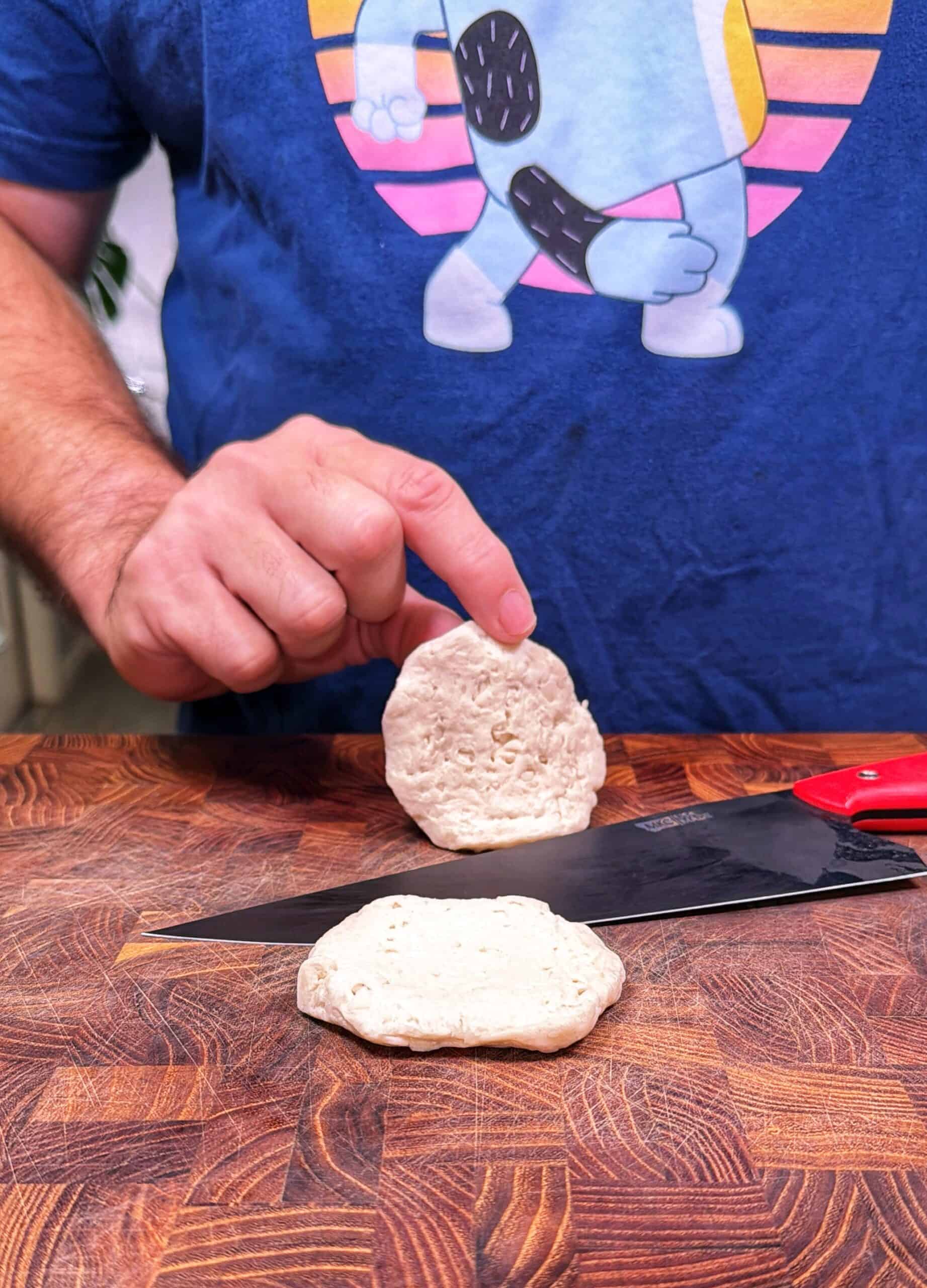 A person wearing a blue shirt with a cartoon robot design is slicing biscuit dough on a wooden <a href=