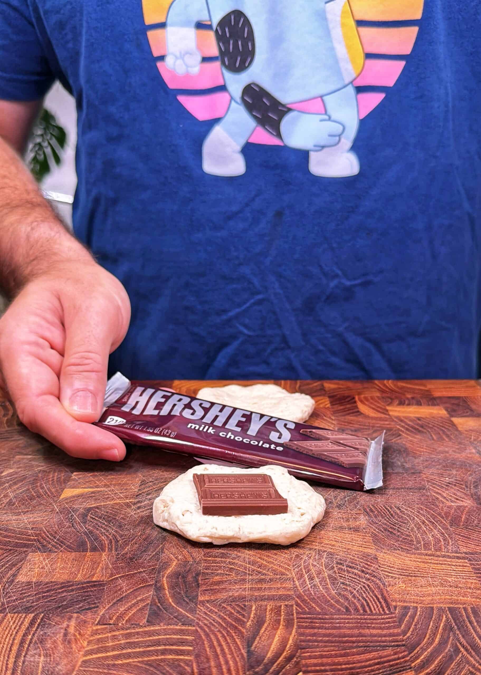 A person wearing a blue t-shirt with a cartoon donut holds a Hersheys milk chocolate bar above a piece of dough with a chocolate square placed on top, all on a wooden surface.