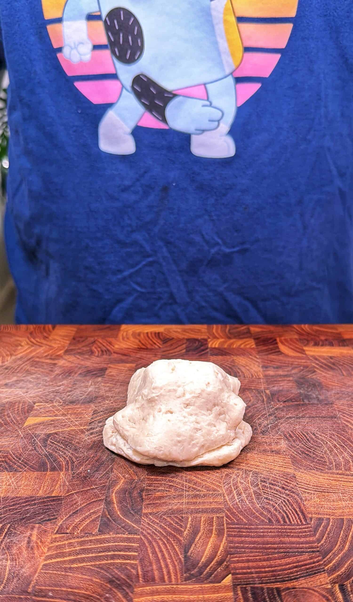 A small, uneven piece of dough rests on a wooden surface, with a person in a blue shirt featuring a cartoon dancing cookie and a sunset design standing in the background.