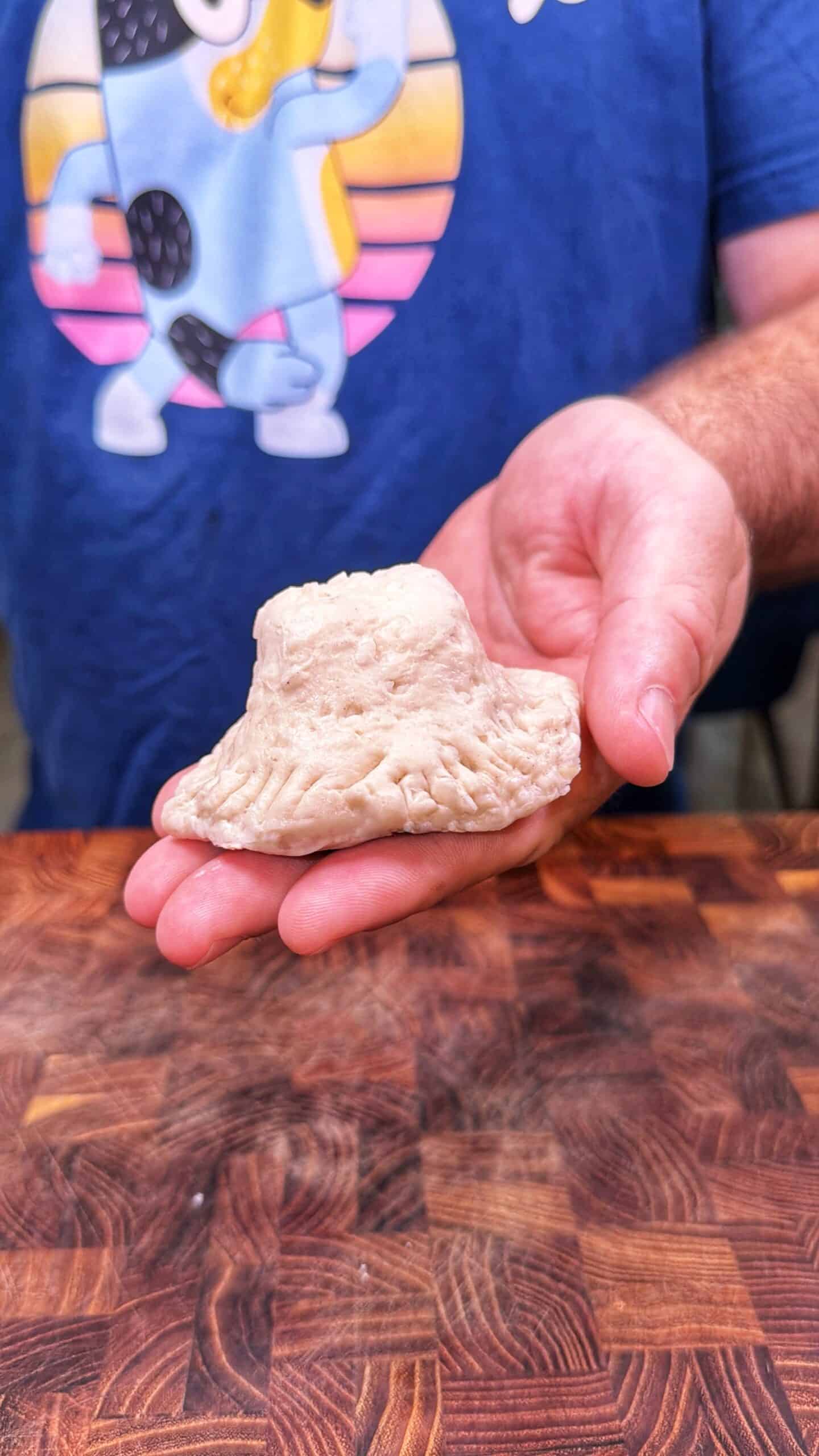 A person holds a piece of dough shaped like a cowboy hat over a wooden surface. A blue shirt with a cartoon character is visible in the background.