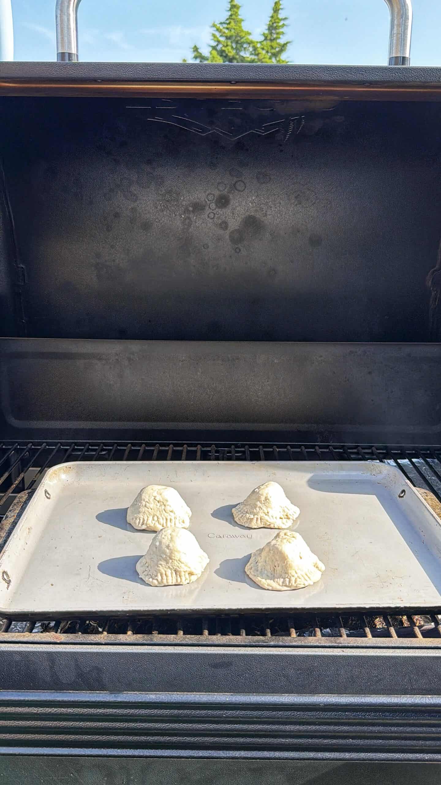 Four uncooked hand pies sit on a baking tray inside an open outdoor grill, with a sunny sky and green tree visible in the background.