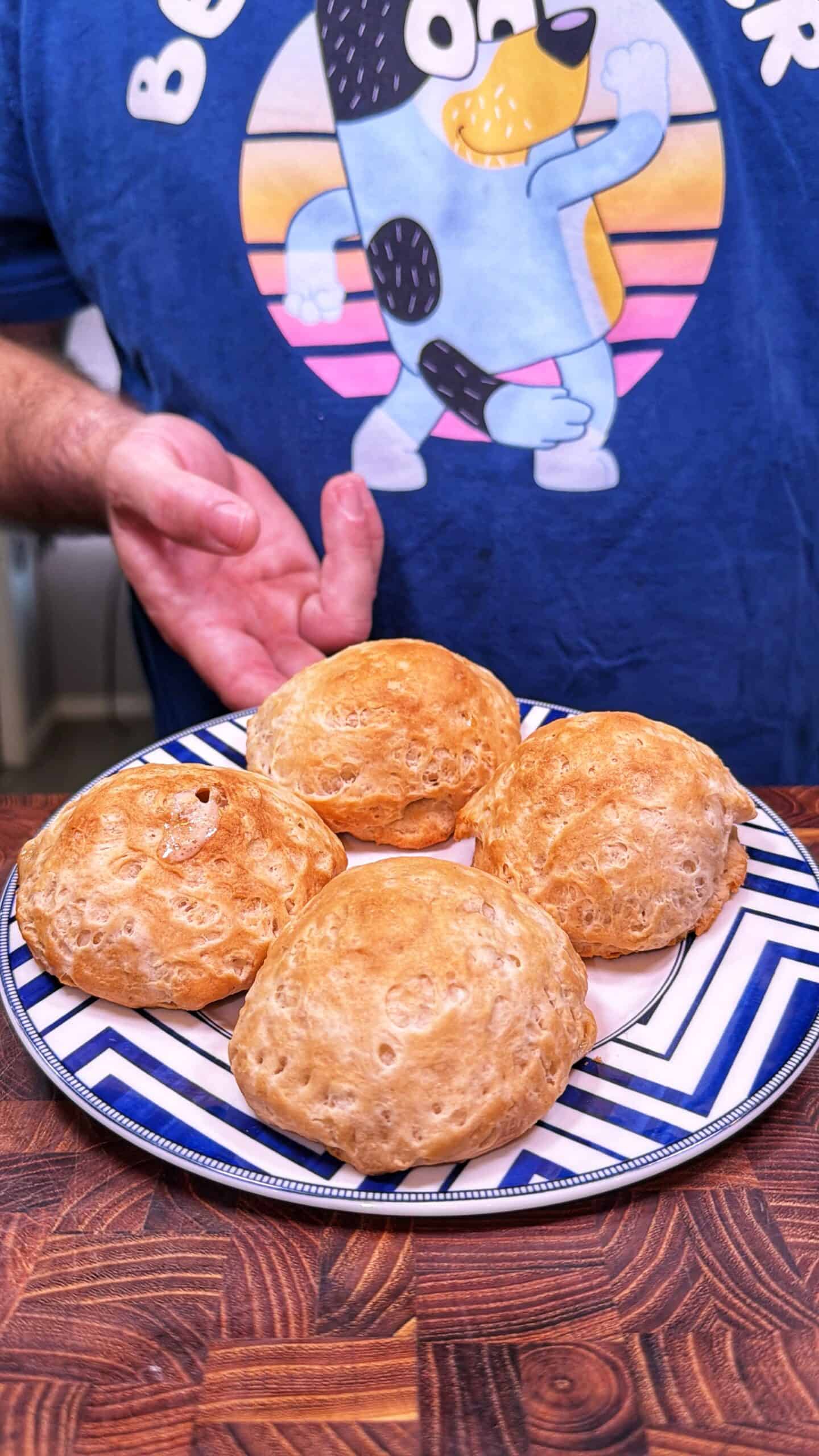 A person wearing a blue t-shirt with a cartoon dog is presenting a plate with four round, golden-brown bread rolls on a patterned blue and white plate, set on a wooden surface.