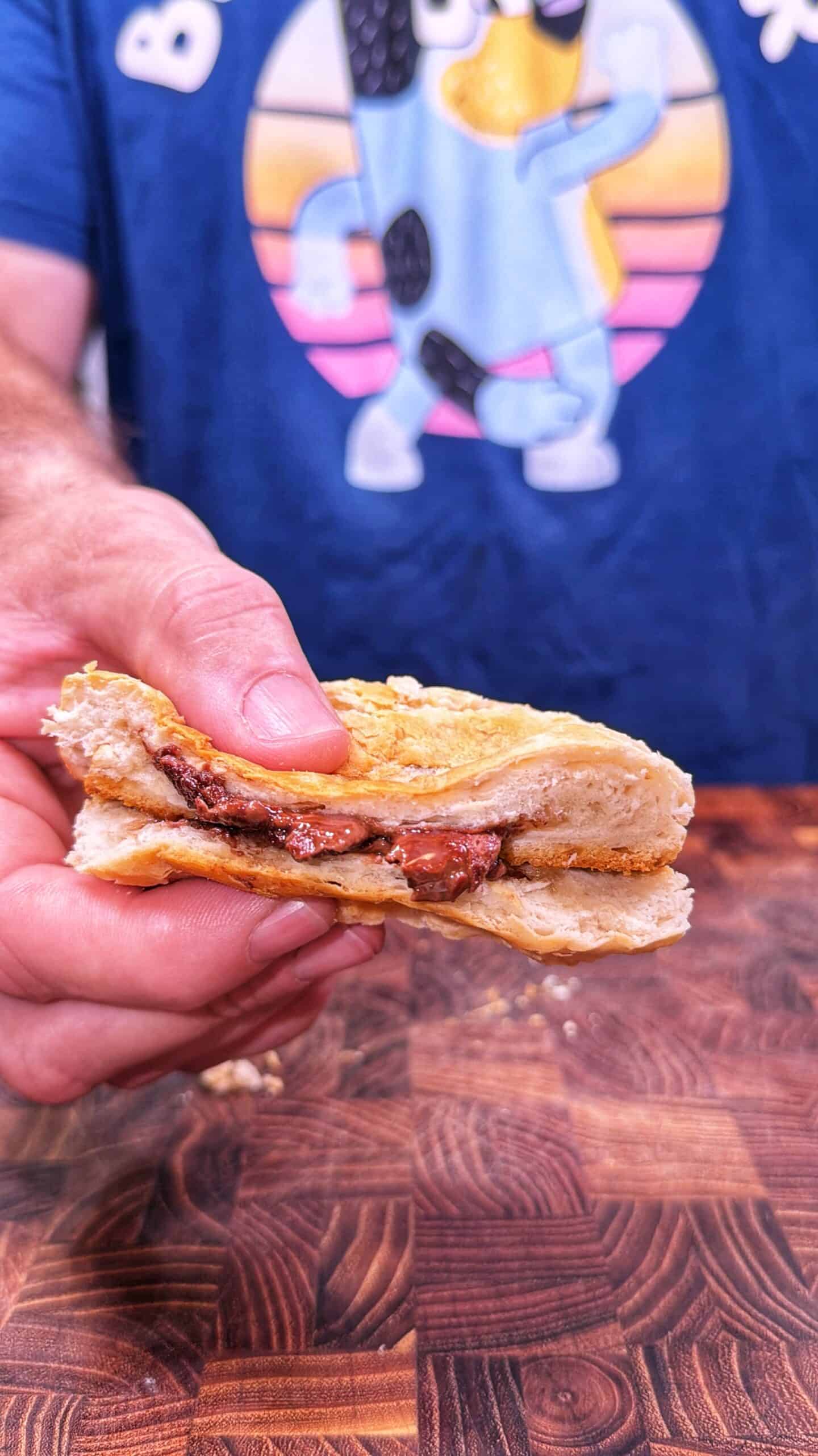 A person holding a toasted sandwich with melted chocolate filling, standing in front of a wooden surface and wearing a blue shirt with a cartoon dog graphic.
