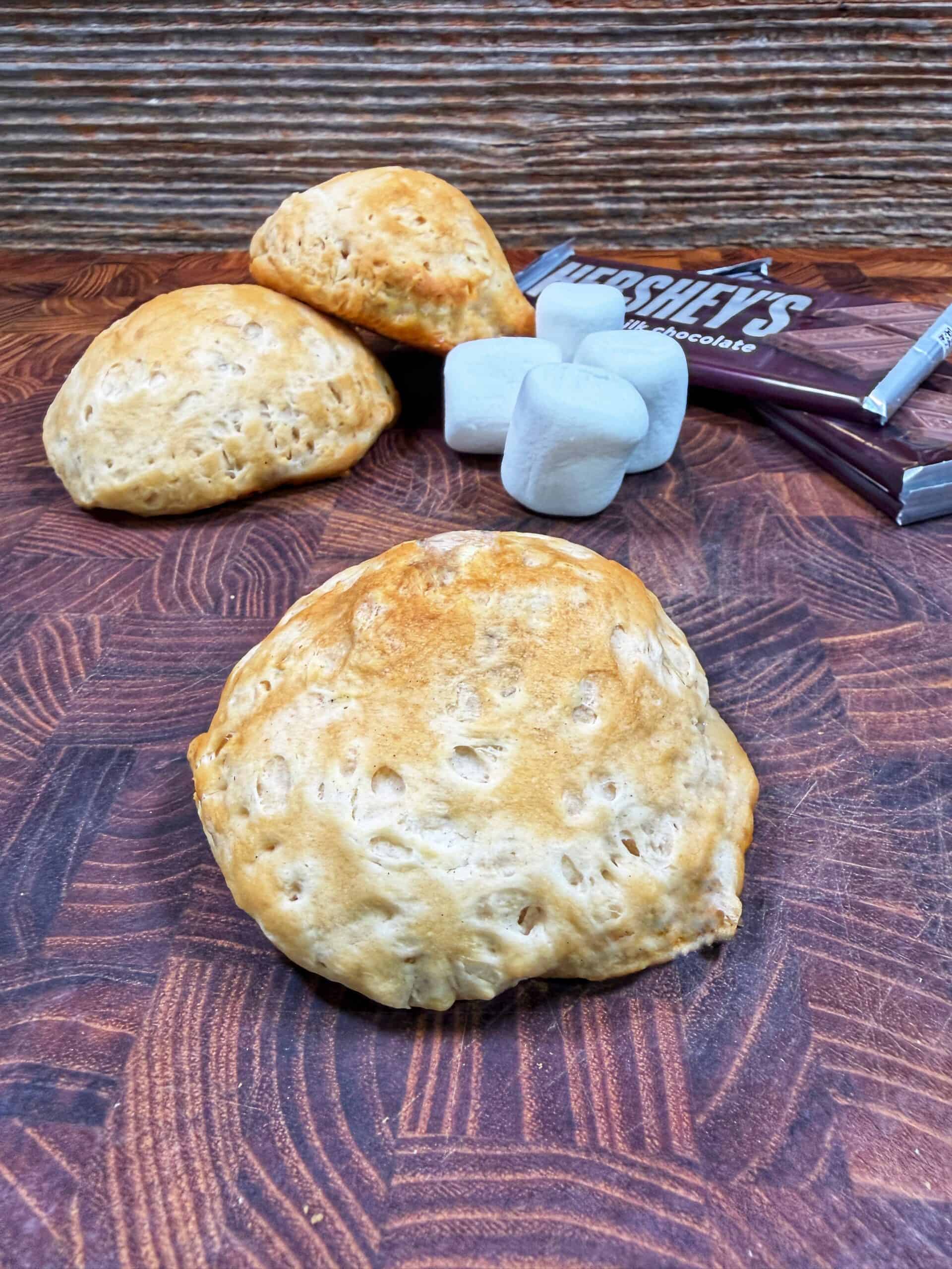 Three pieces of baked bread sit on a wooden surface with a few large marshmallows and Hershey’s chocolate bars in the background. The setting suggests ingredients for making smores.
