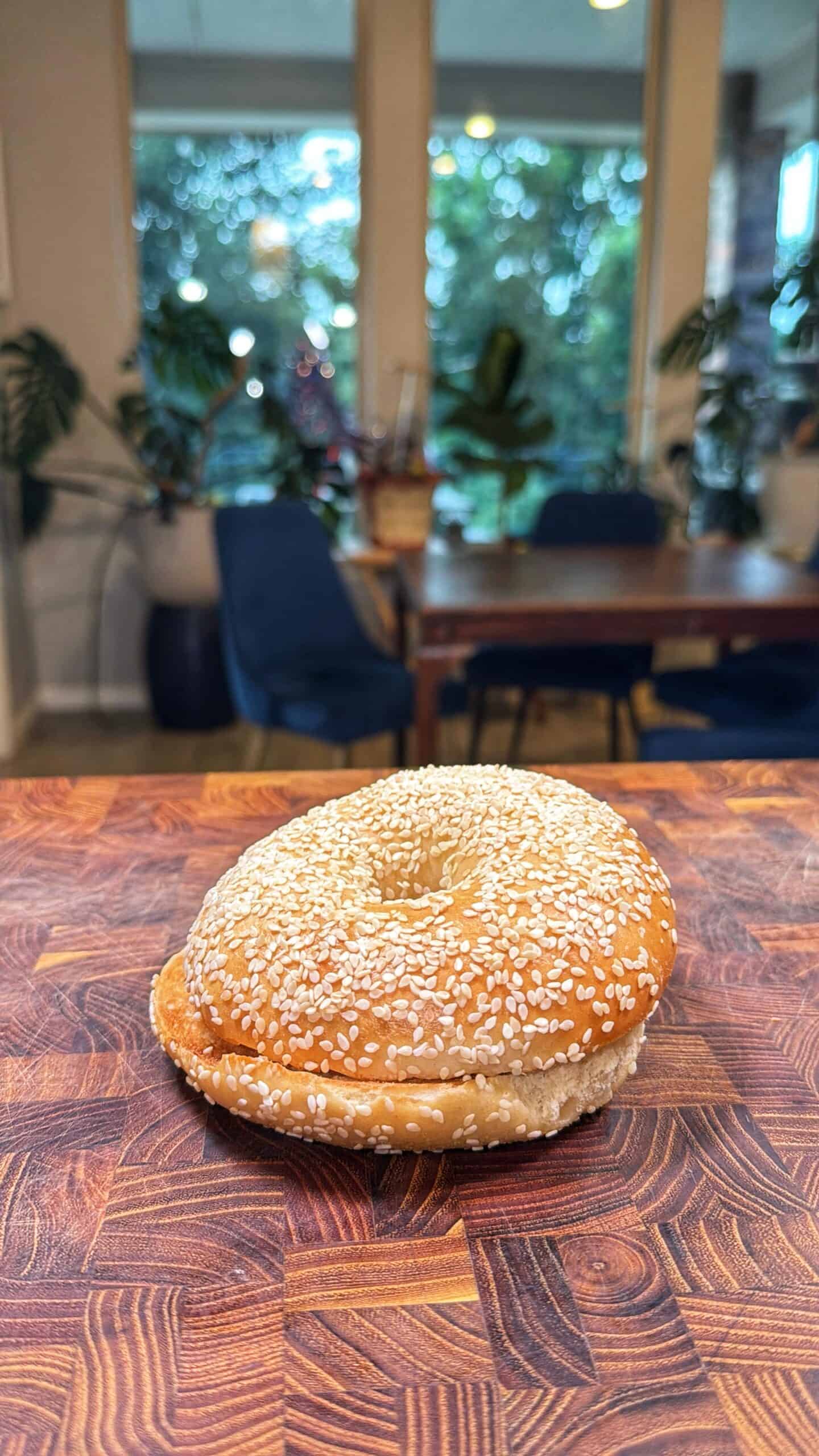 A sesame seed bagel sits on a wooden countertop with a dining table, chairs, large windows, and green plants in the blurred background.