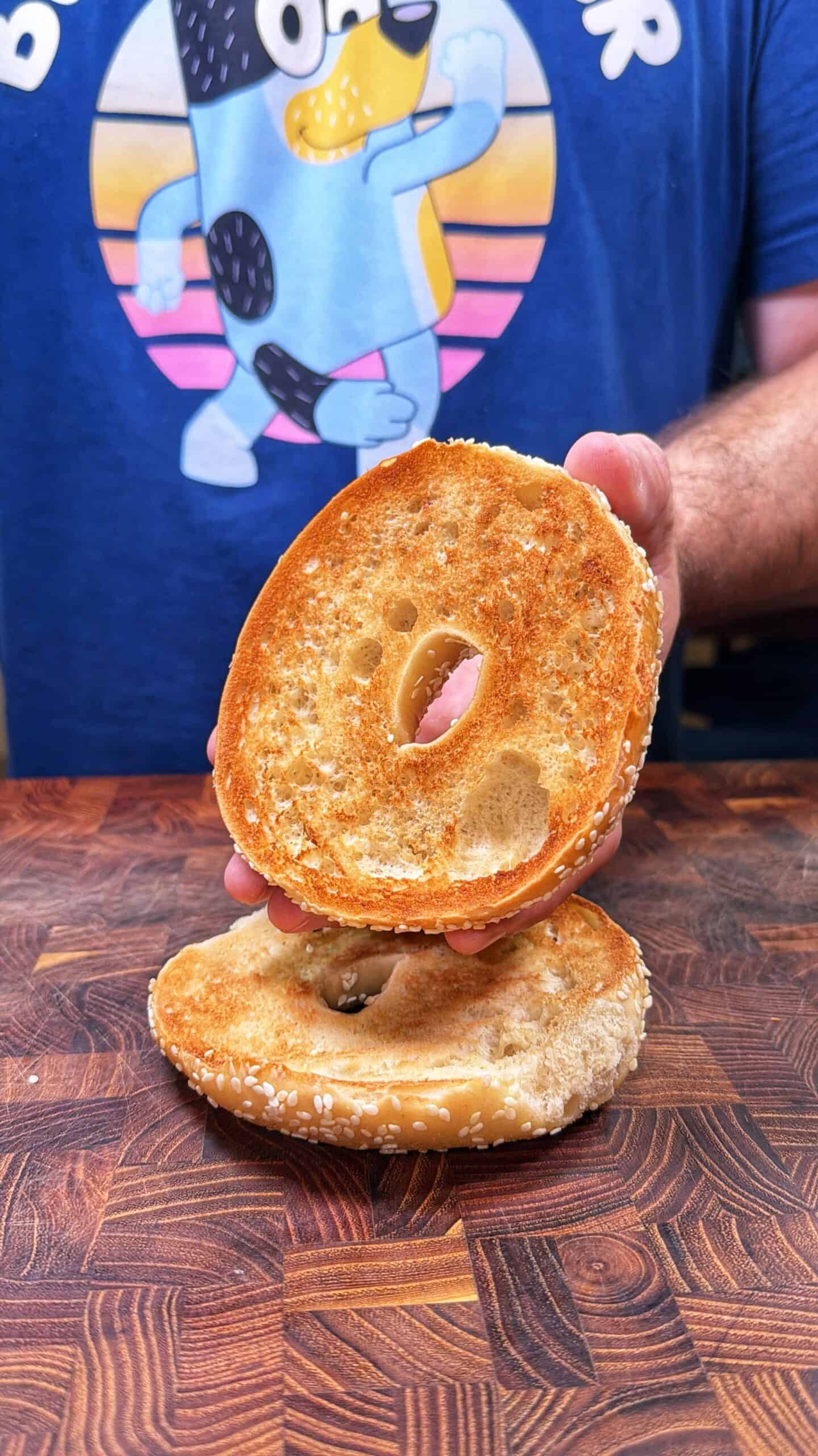 A person holding a toasted sesame bagel half above another half on a wooden surface, wearing a blue shirt with a cartoon dog design.