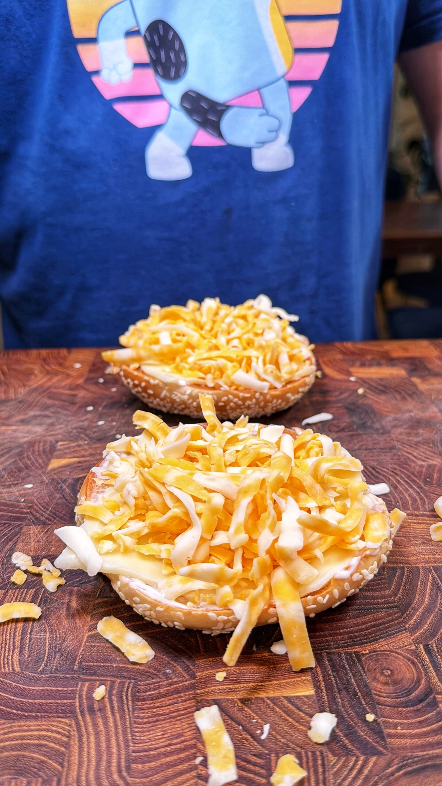 A close-up of two bagel halves topped with a generous amount of shredded cheese on a wooden surface, with a person in a blue shirt featuring a cartoon character in the background.