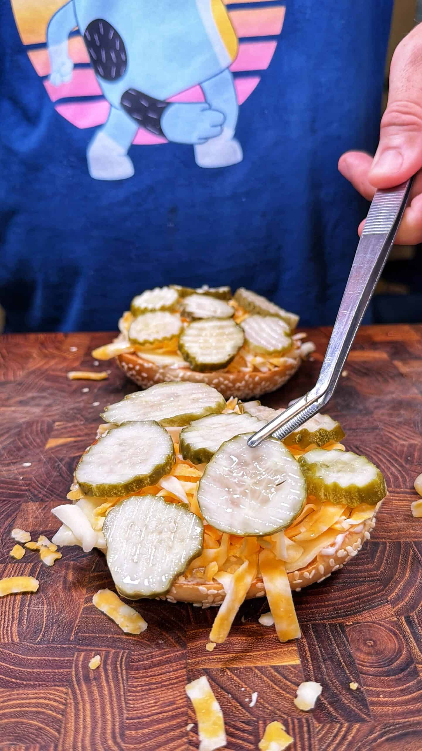 A person uses tongs to place pickle slices on a sandwich bun topped with shredded cheese and onions. Another bun with similar toppings is in the background. The person is wearing a blue shirt with a cartoon character design.