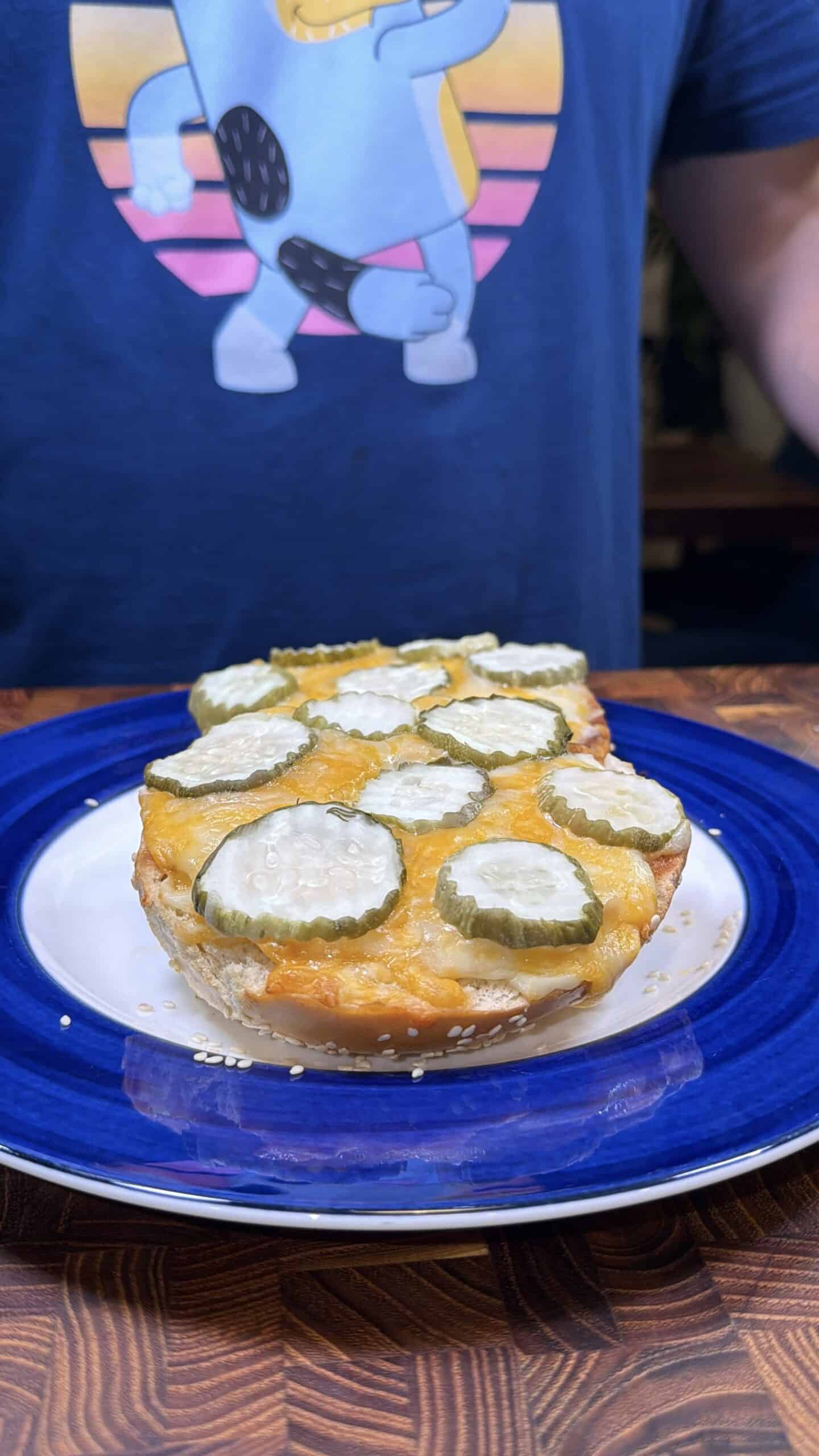 A sesame bun half topped with melted cheese and sliced pickles sits on a white and blue plate. In the background, a person wearing a blue T-shirt with a cartoon graphic stands behind the plate.