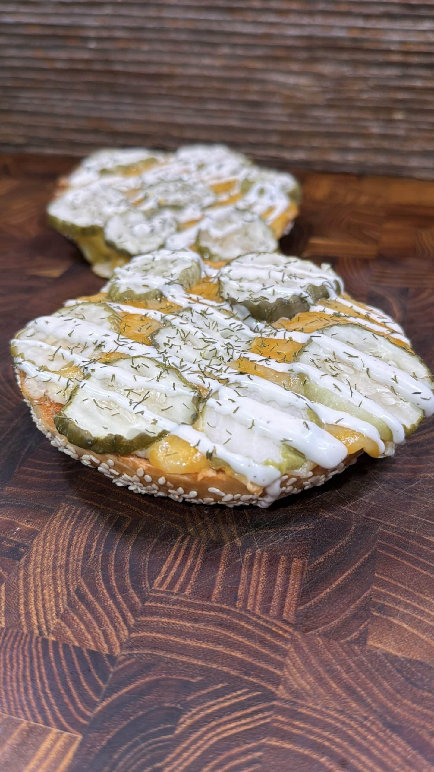 A close-up of a sliced bagel topped with melted cheese, pickles, creamy white sauce, and a sprinkle of dried herbs on a wooden surface. Another similarly topped bagel slice is in the background.