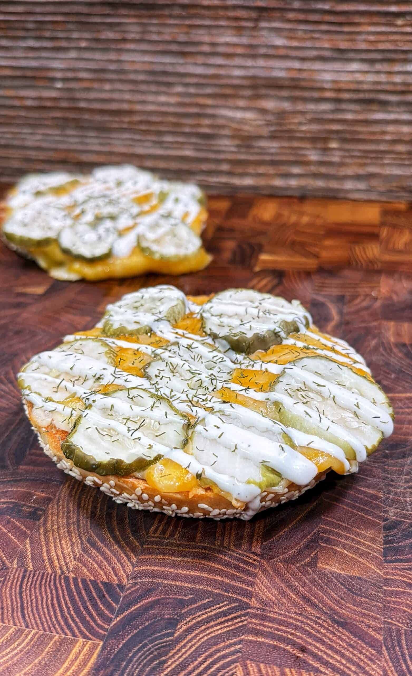 A round, open-faced sandwich topped with sliced pickles, melted cheese, white sauce, and sprinkled herbs sits on a wooden surface, with another similar sandwich blurred in the background.