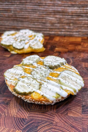 A round sandwich bun topped with sliced pickles, melted cheese, a drizzle of white sauce, and sprinkled herbs sits on a patterned wooden surface; another similar item is blurred in the background.