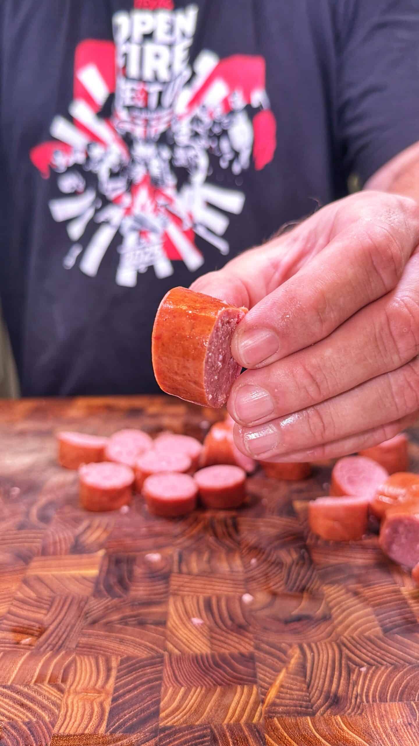 A person holds a sliced piece of sausage over a wooden cutting board with more sausage slices. The person wears a black T-shirt with a white and red graphic design.