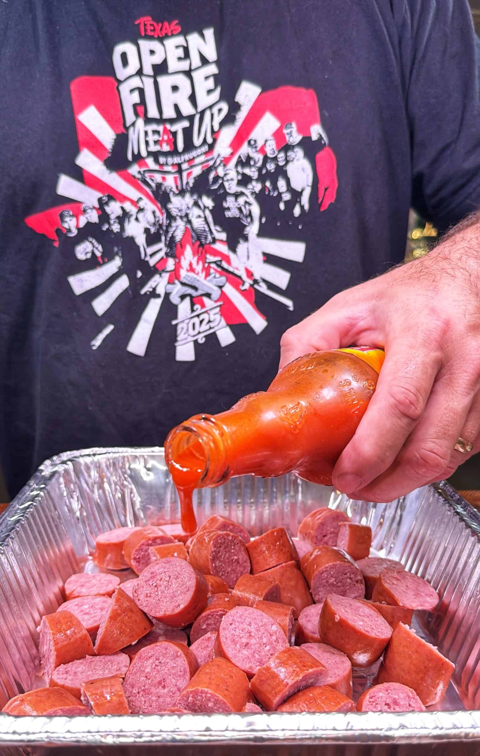 A person in an “Open Fire Meat Up” t-shirt pours hot sauce over sliced sausage pieces in a foil tray.