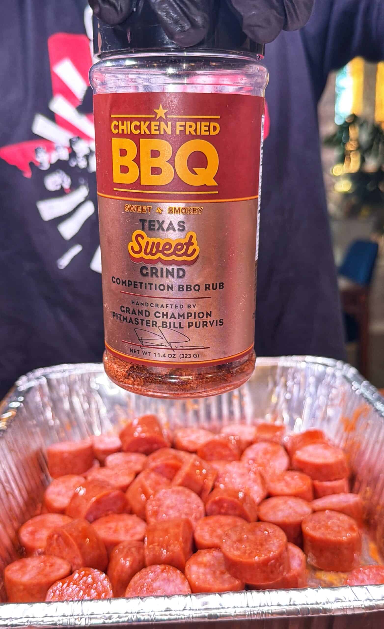 A gloved hand holds a jar of Texas Sweet Grind Chicken Fried BBQ rub above a foil tray filled with sliced sausages. The jar’s label is prominently displayed in the foreground.
