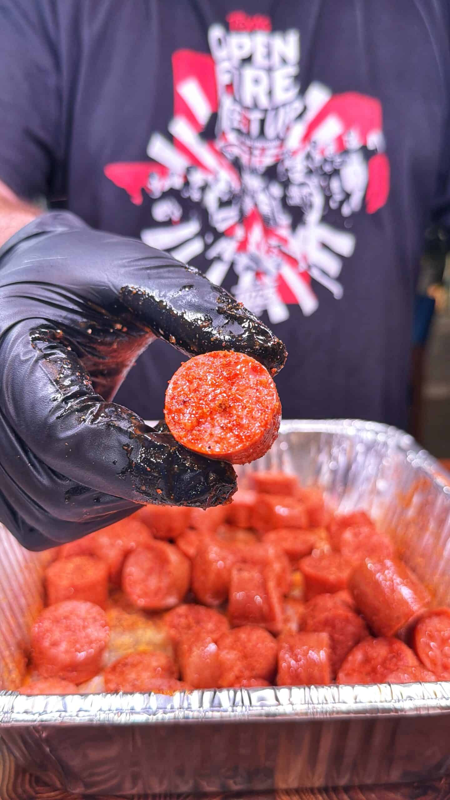 A person wearing a black glove holds up a sliced piece of sausage over a tray filled with more sausage pieces. The person is wearing a black shirt with a red and white graphic design.