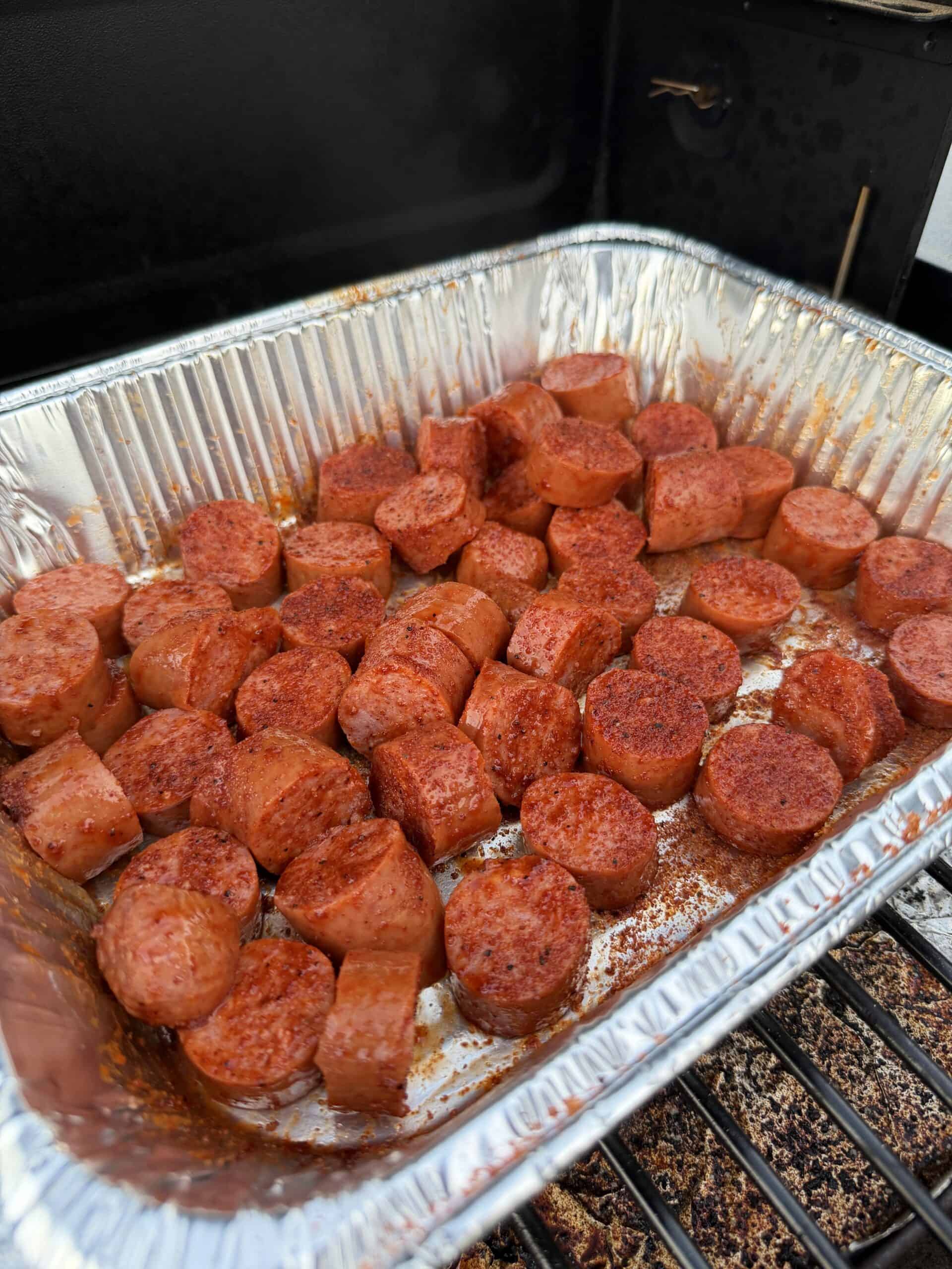 Sliced sausage pieces seasoned with spices are arranged in a disposable aluminum tray on a grill, ready to be cooked.