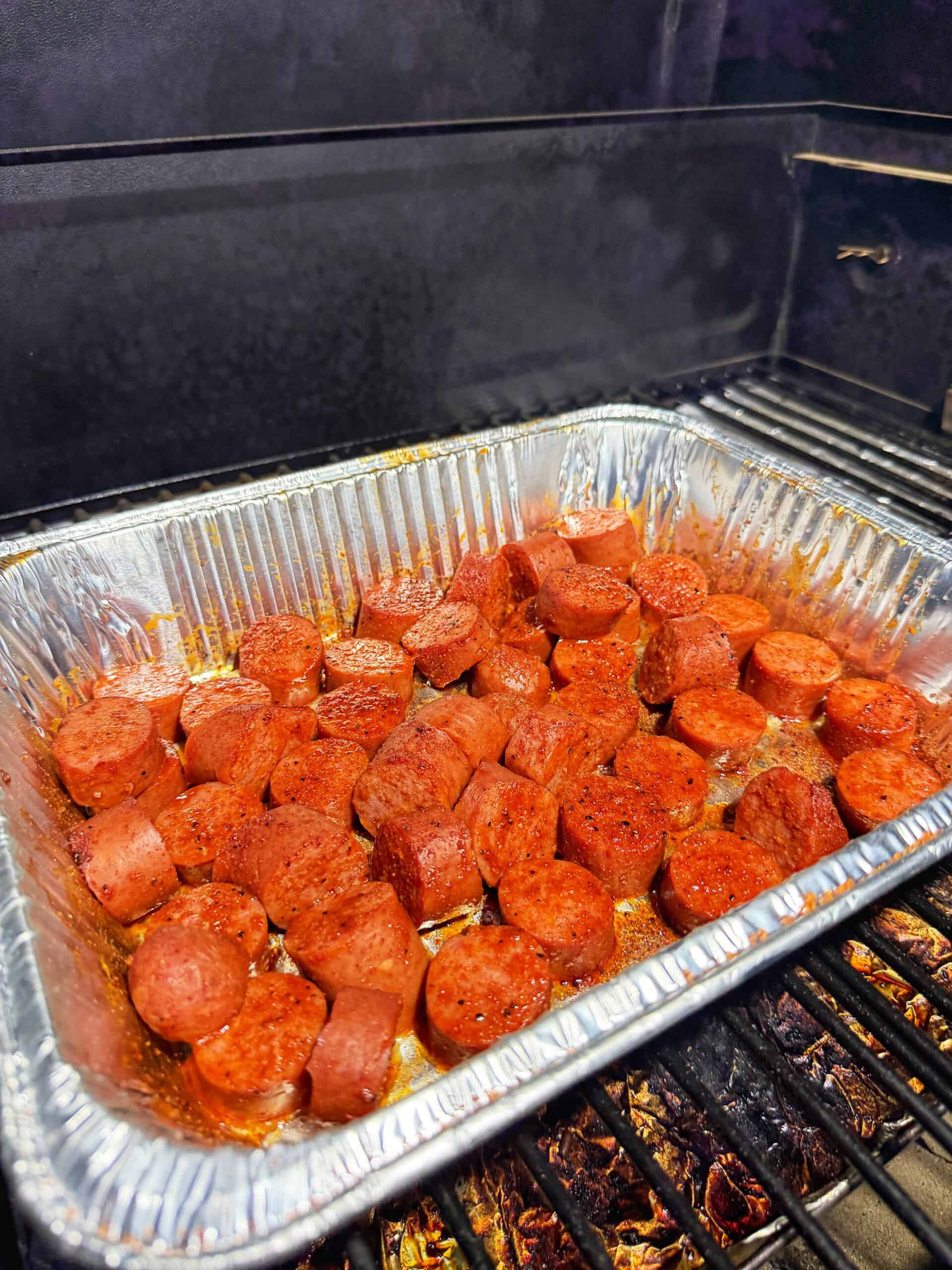Sliced sausage pieces are seasoned and arranged in a disposable aluminum tray on a grill, being cooked.