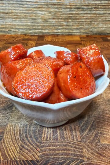 A white bowl filled with round slices of glazed, cooked sausage sits on a wooden surface with a rustic, wood-textured background.