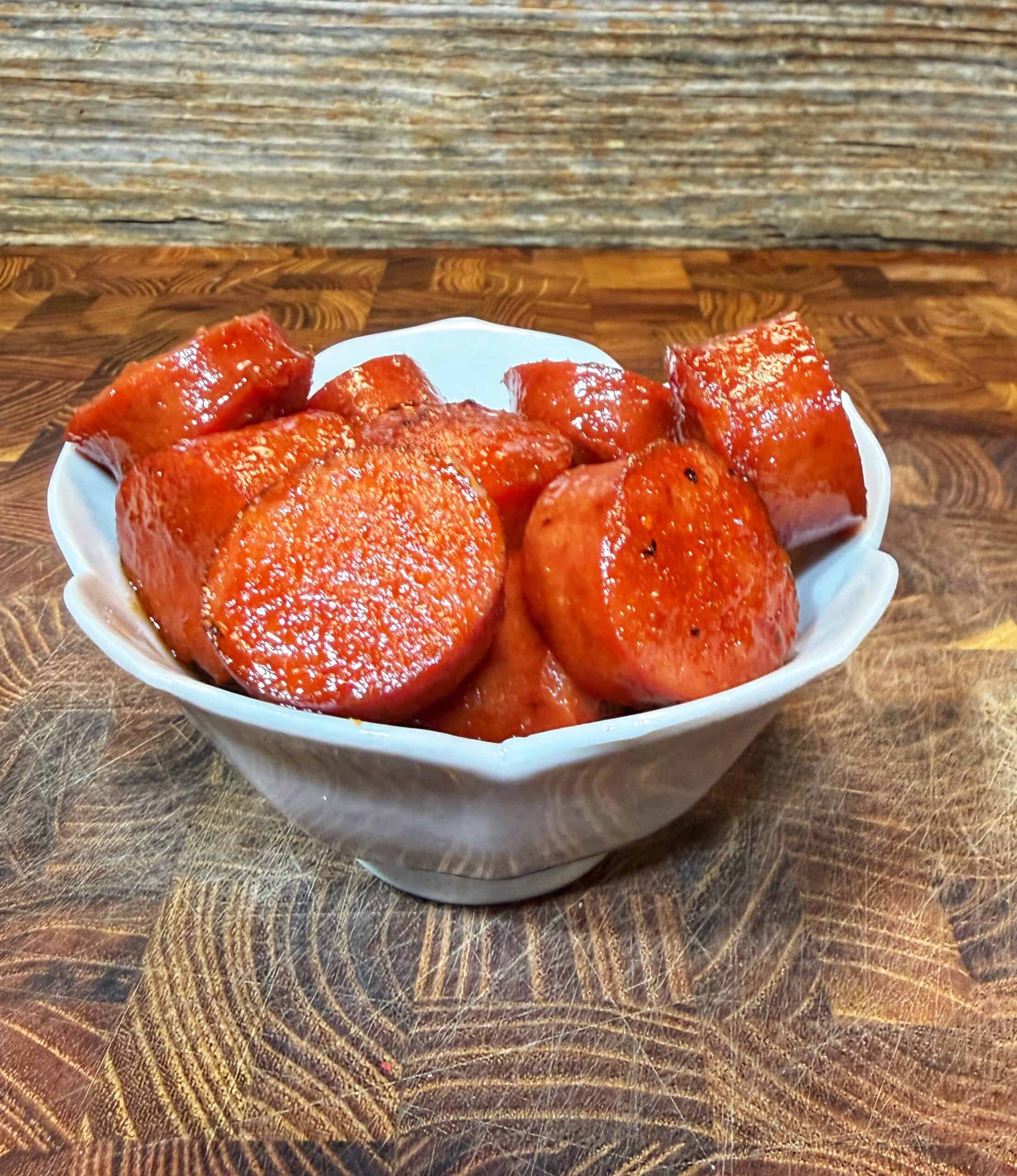 A white bowl filled with round slices of glazed, cooked sausage sits on a wooden surface with a rustic, wood-textured background.