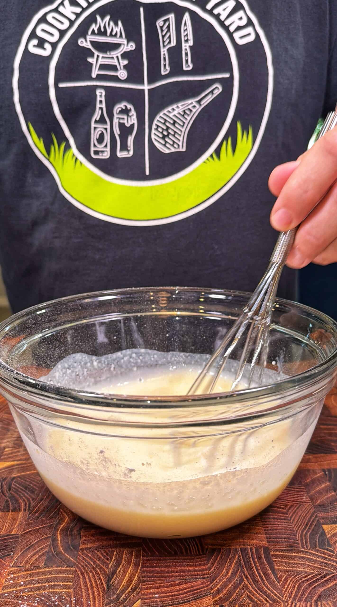 A person whisking a light brown mixture in a glass bowl on a wooden surface, wearing a shirt with outdoor cooking graphics.