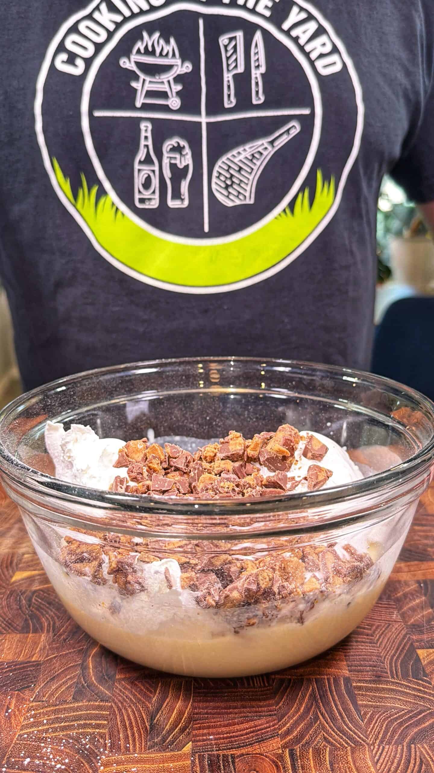 A clear glass bowl holds a mixture of batter, whipped topping, and chopped chocolate bars on a wooden surface. In the background, a person wears a Cooking in the Yard t-shirt.