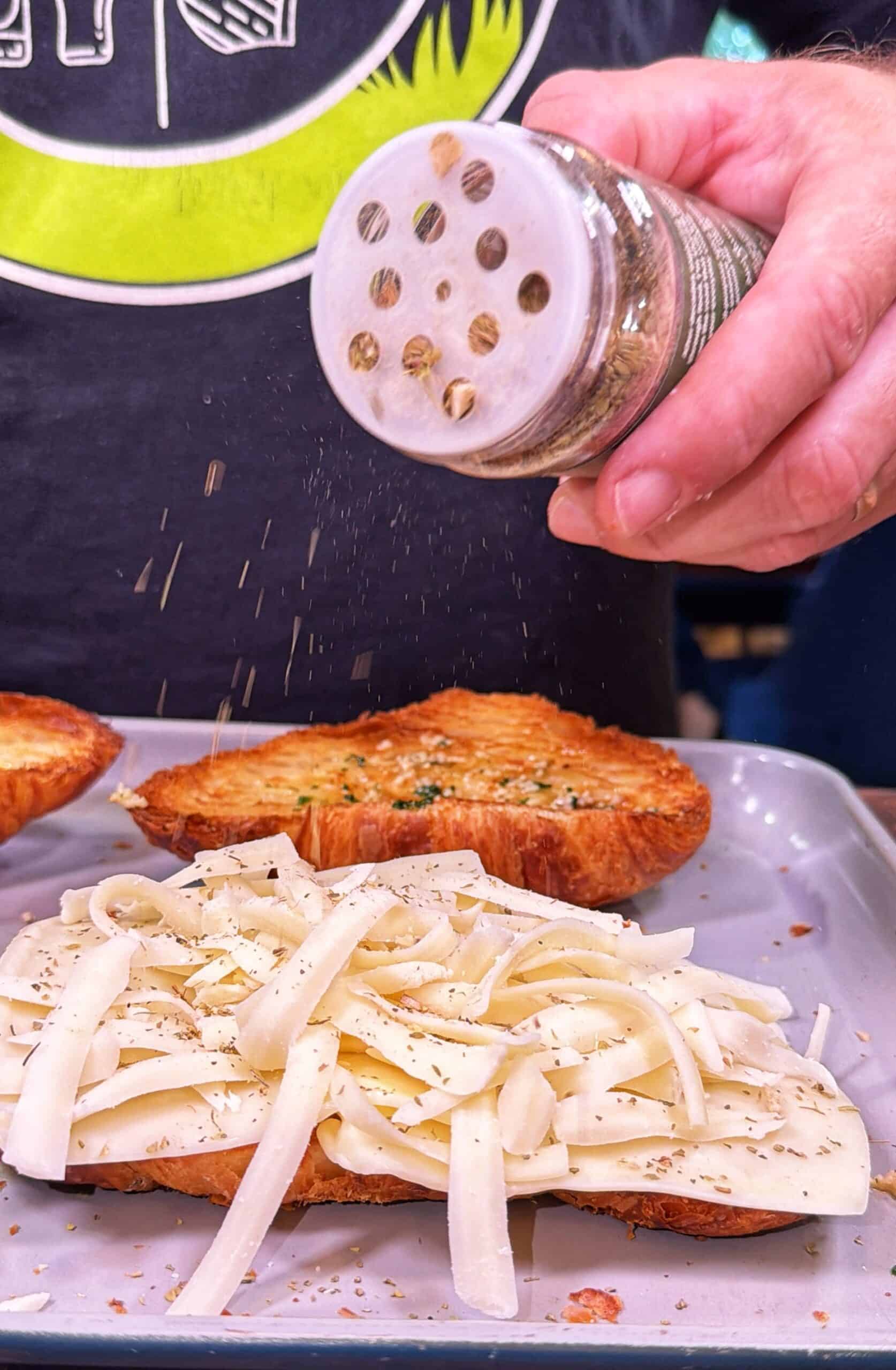 A hand sprinkles seasoning onto bread topped with shredded cheese, with another slice of bread on a tray nearby.