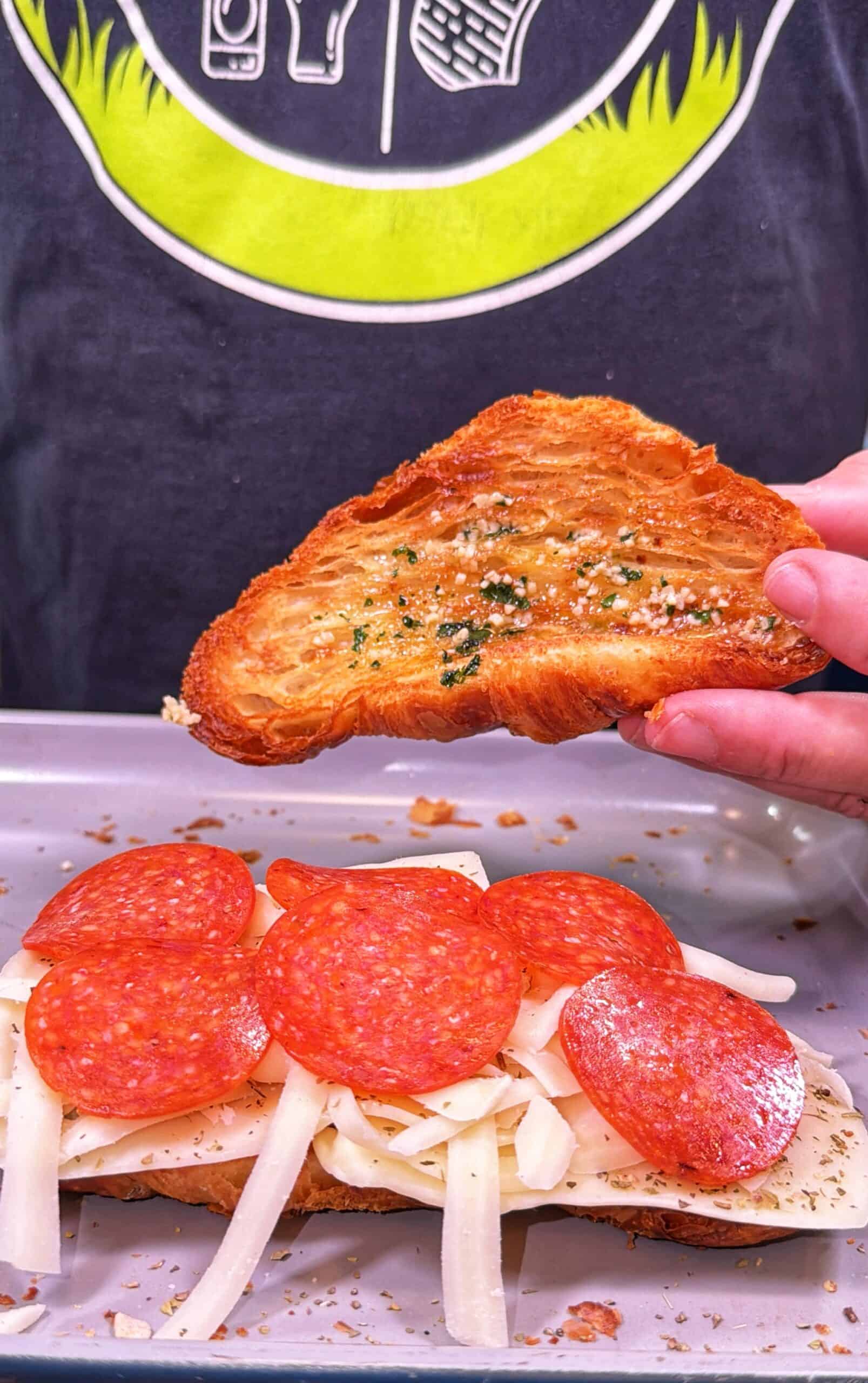 A person holds a slice of toasted, seasoned bread above a sandwich topped with mozzarella cheese and pepperoni slices on a tray.