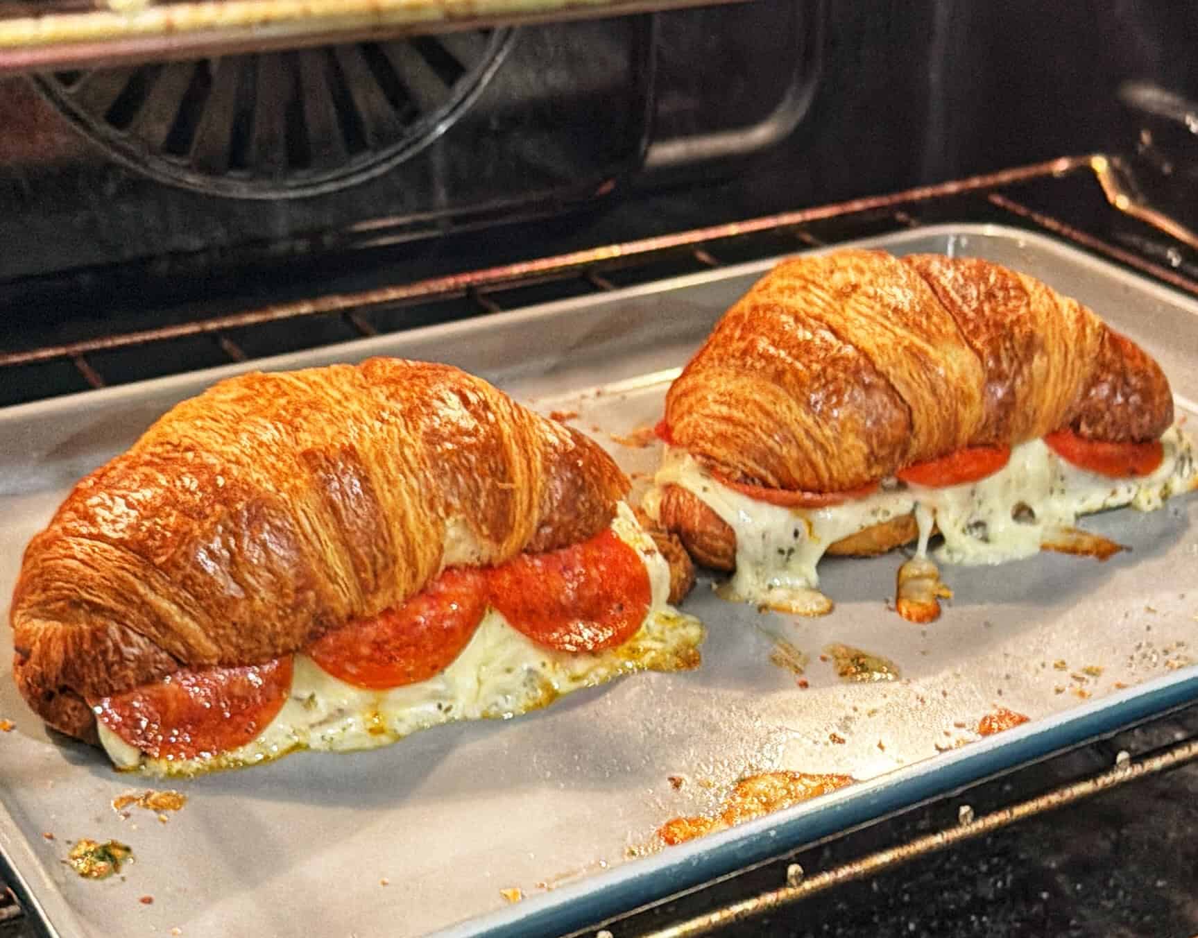 Two croissant sandwiches on a baking tray in an oven, filled with melted cheese and pepperoni slices. The cheese is slightly oozing out, and the croissants are golden brown.