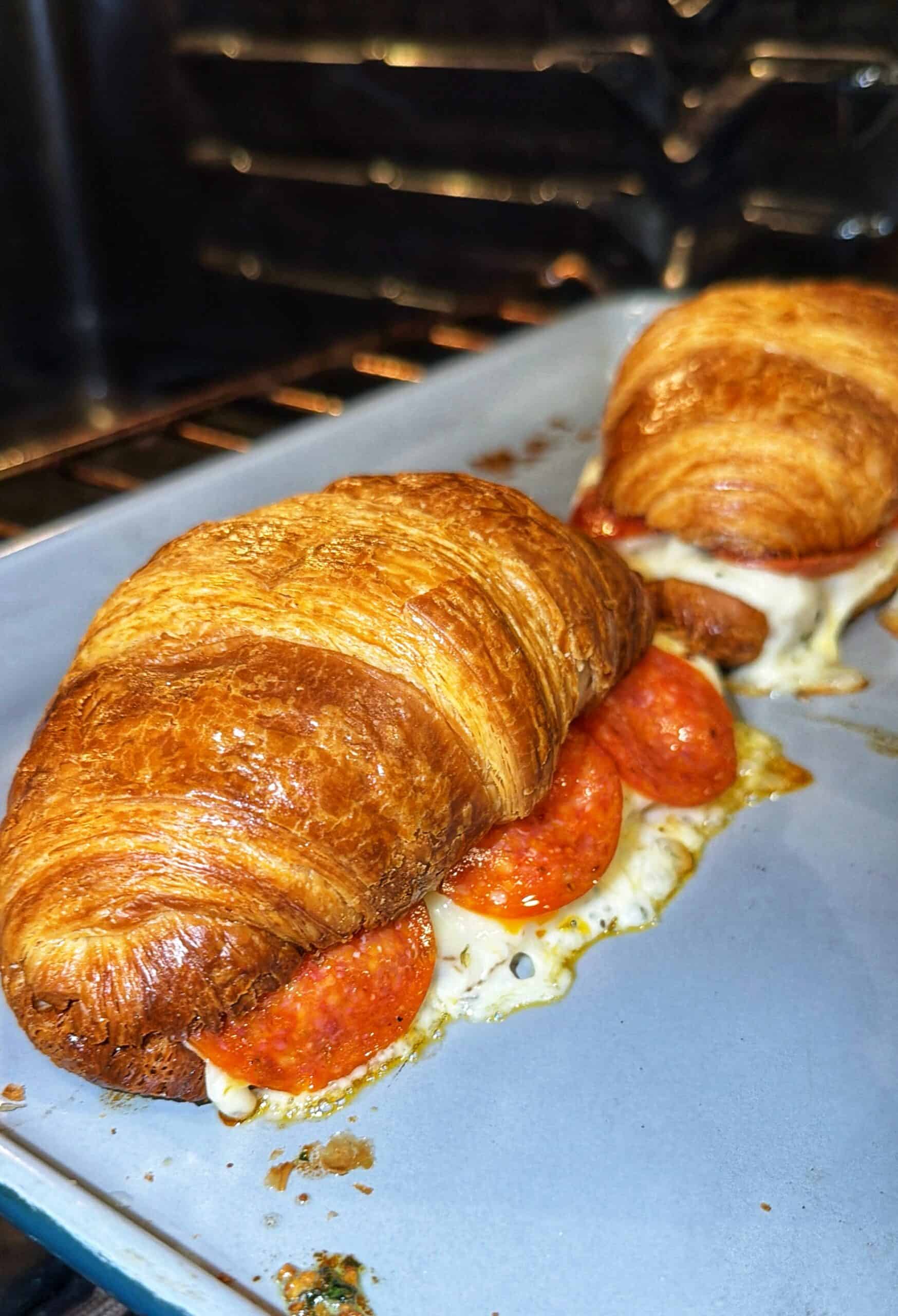 Two croissants filled with melted cheese and pepperoni slices are baking on a tray in the oven.