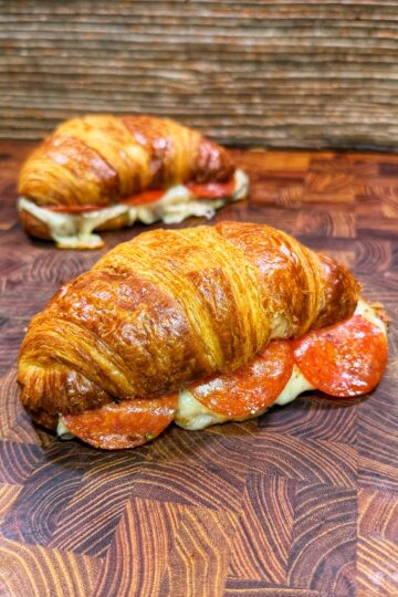 Two golden croissant sandwiches with melted cheese and pepperoni slices on a patterned wooden surface, one in the foreground and one in the background.