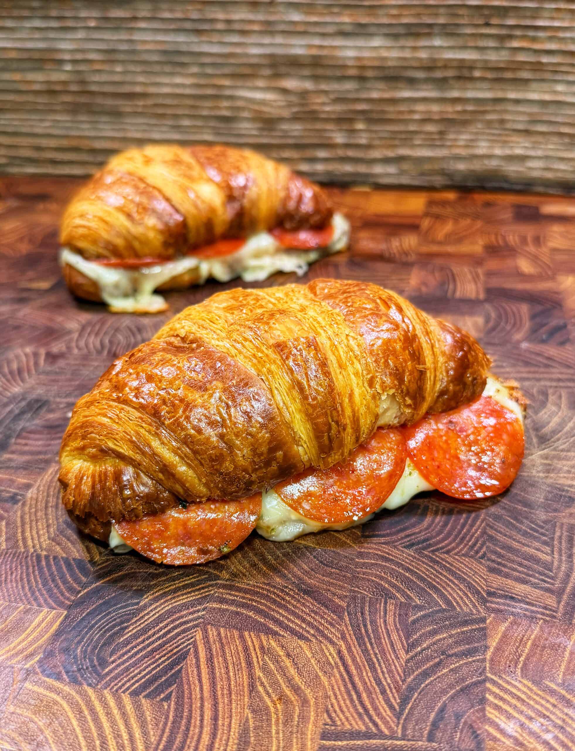 Two golden croissant sandwiches with melted cheese and pepperoni slices on a patterned wooden surface, one in the foreground and one in the background.