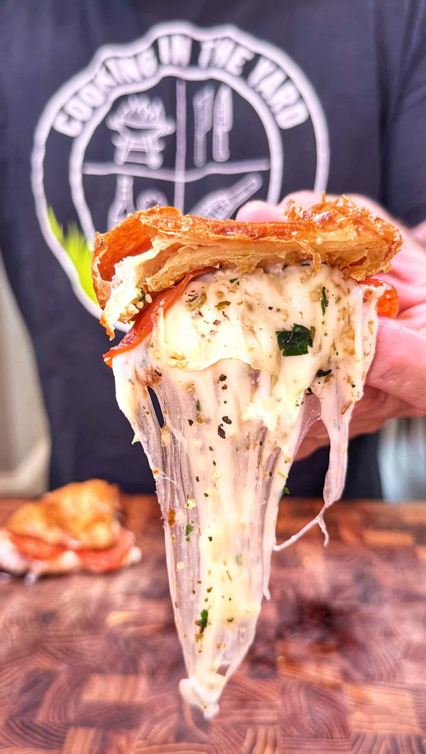 A hand lifts a cheesy slice of pizza with herbs and pepperoni, cheese stretching as its pulled away from the rest on a wooden surface. A person in a dark shirt with a circular logo is in the background.