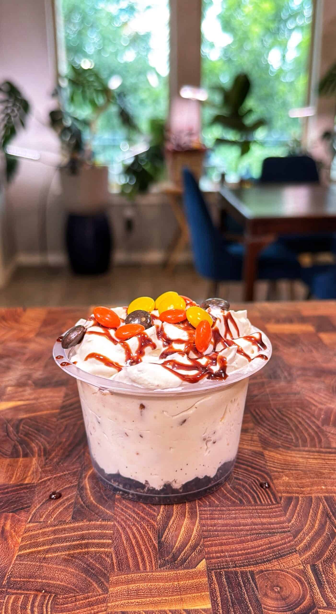 A cup of creamy dessert with chocolate chips, whipped topping, drizzled sauce, and colorful candies sits on a wooden table in a bright, plant-filled room.