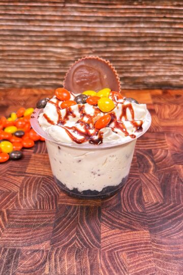 A cup of creamy dessert topped with whipped cream, chocolate drizzle, orange and yellow candy-coated chocolates, and a chocolate peanut butter cup, on a wooden surface with more candies beside it.