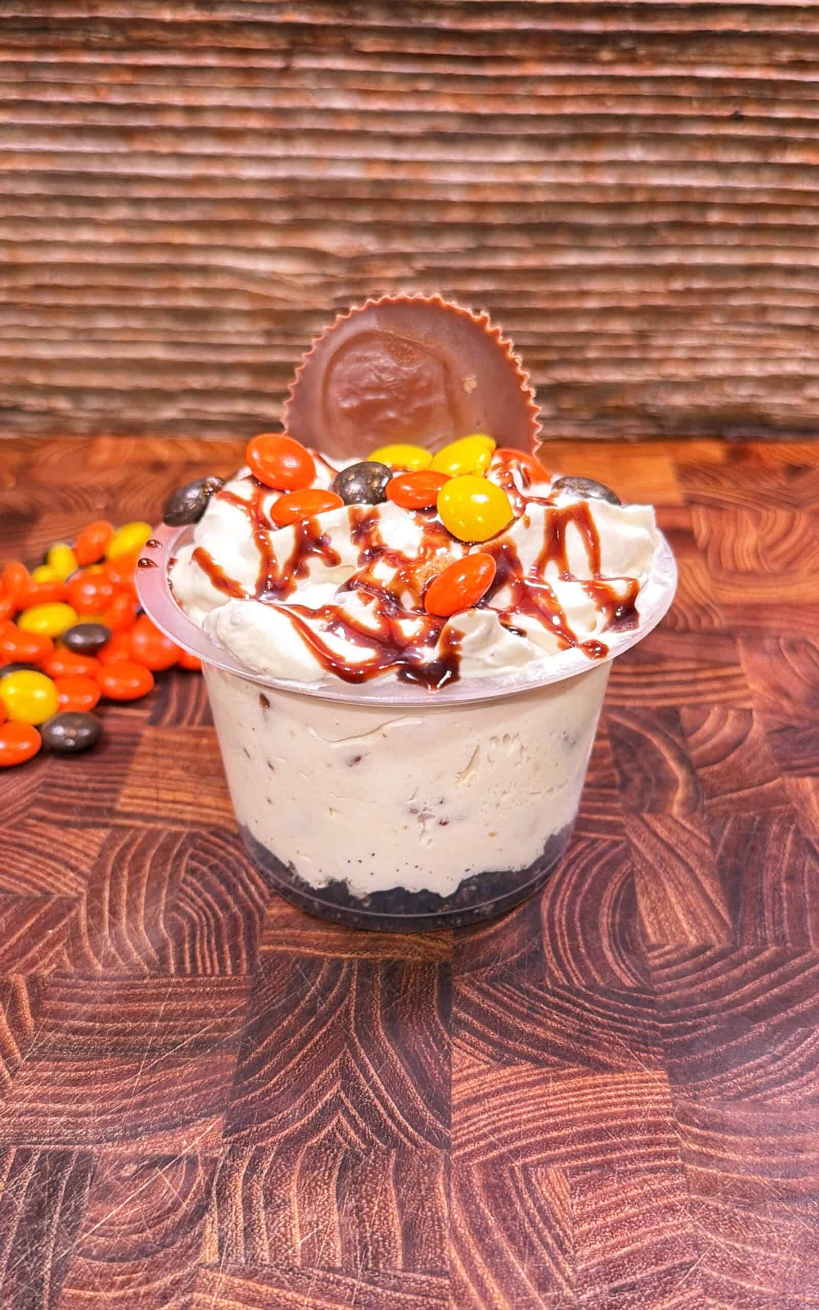 A cup of creamy dessert topped with whipped cream, chocolate drizzle, orange and yellow candy-coated chocolates, and a chocolate peanut butter cup, on a wooden surface with more candies beside it.