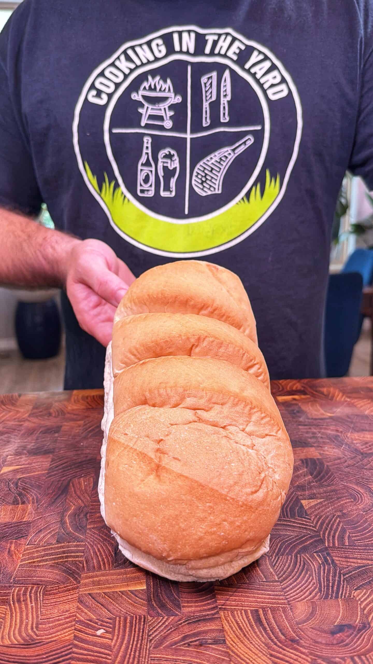 A person in a Cooking in the Yard shirt holds a pack of four sandwich buns on a wooden cutting board.