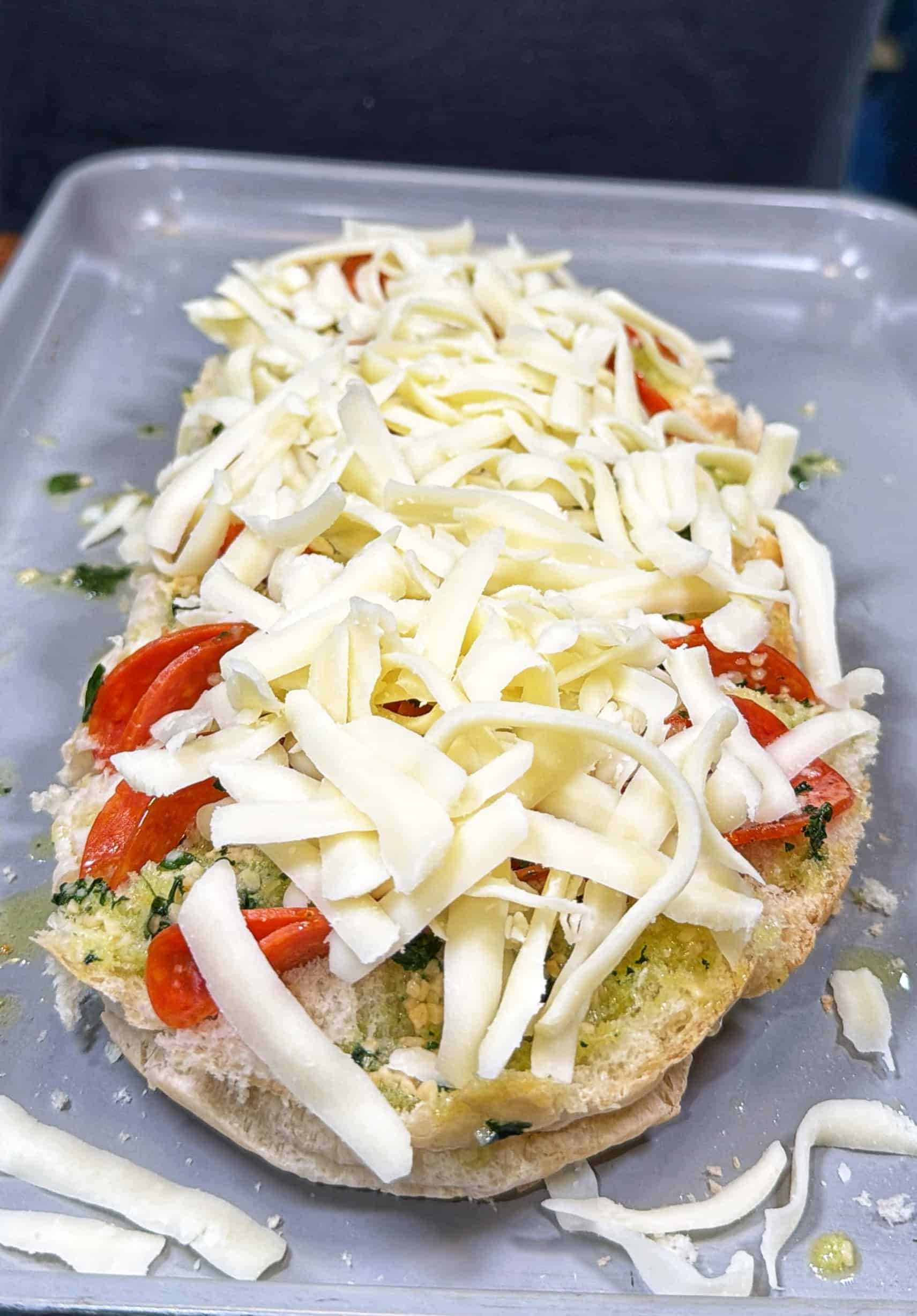 A piece of bread topped with sliced tomatoes, herbs, and a generous amount of shredded cheese sits on a gray baking tray, ready to be baked.